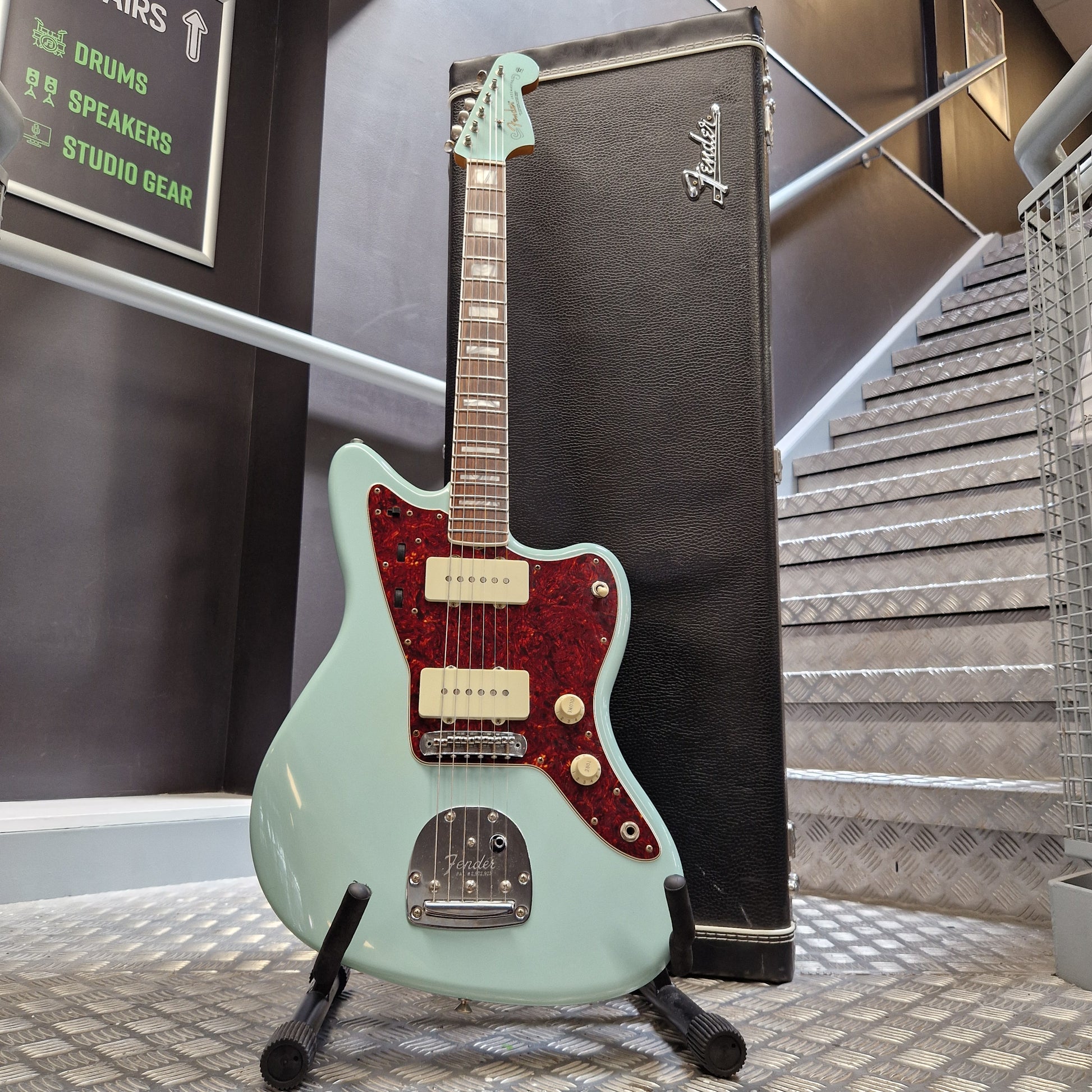 Pre-Owned Fender A mint green Fender Stratocaster electric guitar with a red glitter pickguard, resting on a black Fender amp head against a textured metal wall.
