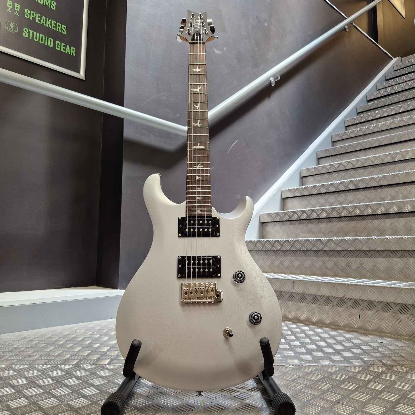 Pre-Owned PRS SE CE24 A white electric guitar with black hardware and gold-toned pickups, displayed on a stand in an indoor setting with metal stair railings and textured walls.