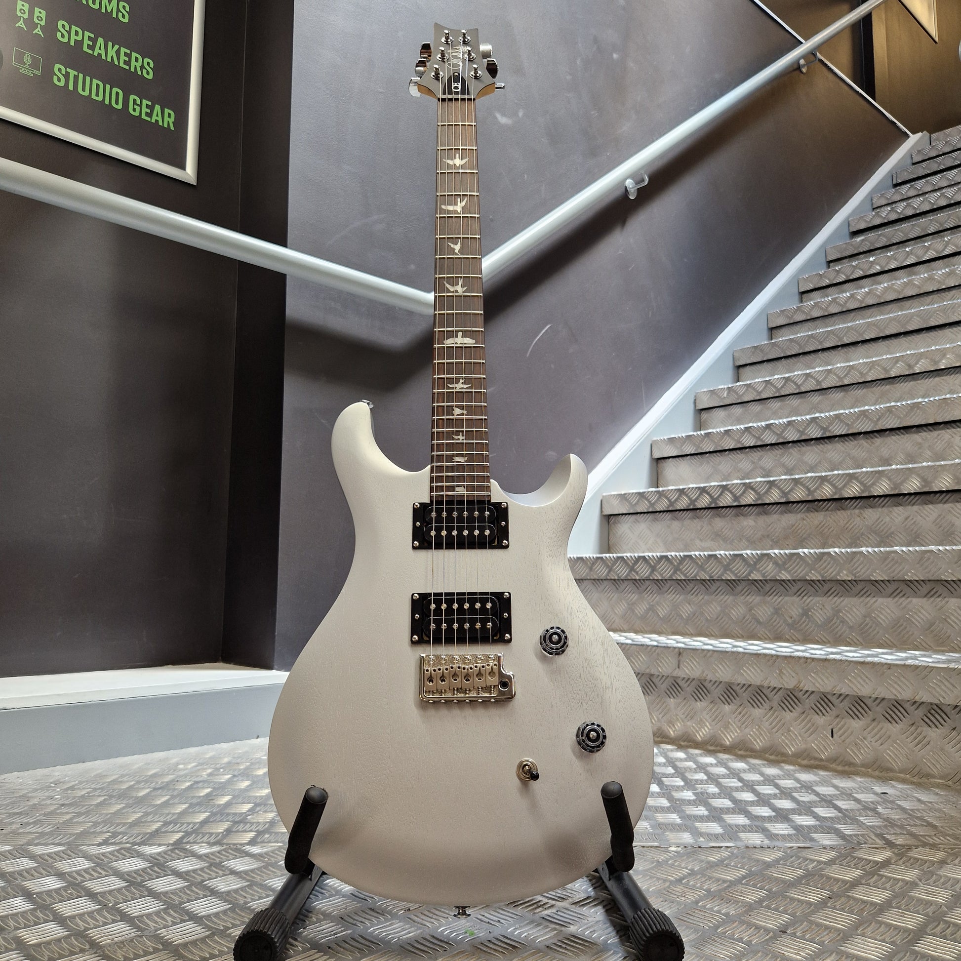 Pre-Owned PRS SE CE24 A white electric guitar with black hardware and gold-toned pickups, displayed on a stand in an indoor setting with metal stair railings and textured walls.