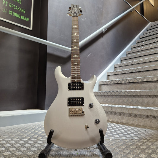 Pre-Owned PRS SE CE24 A white electric guitar with black hardware and gold-toned pickups, displayed on a stand in an indoor setting with metal stair railings and textured walls.