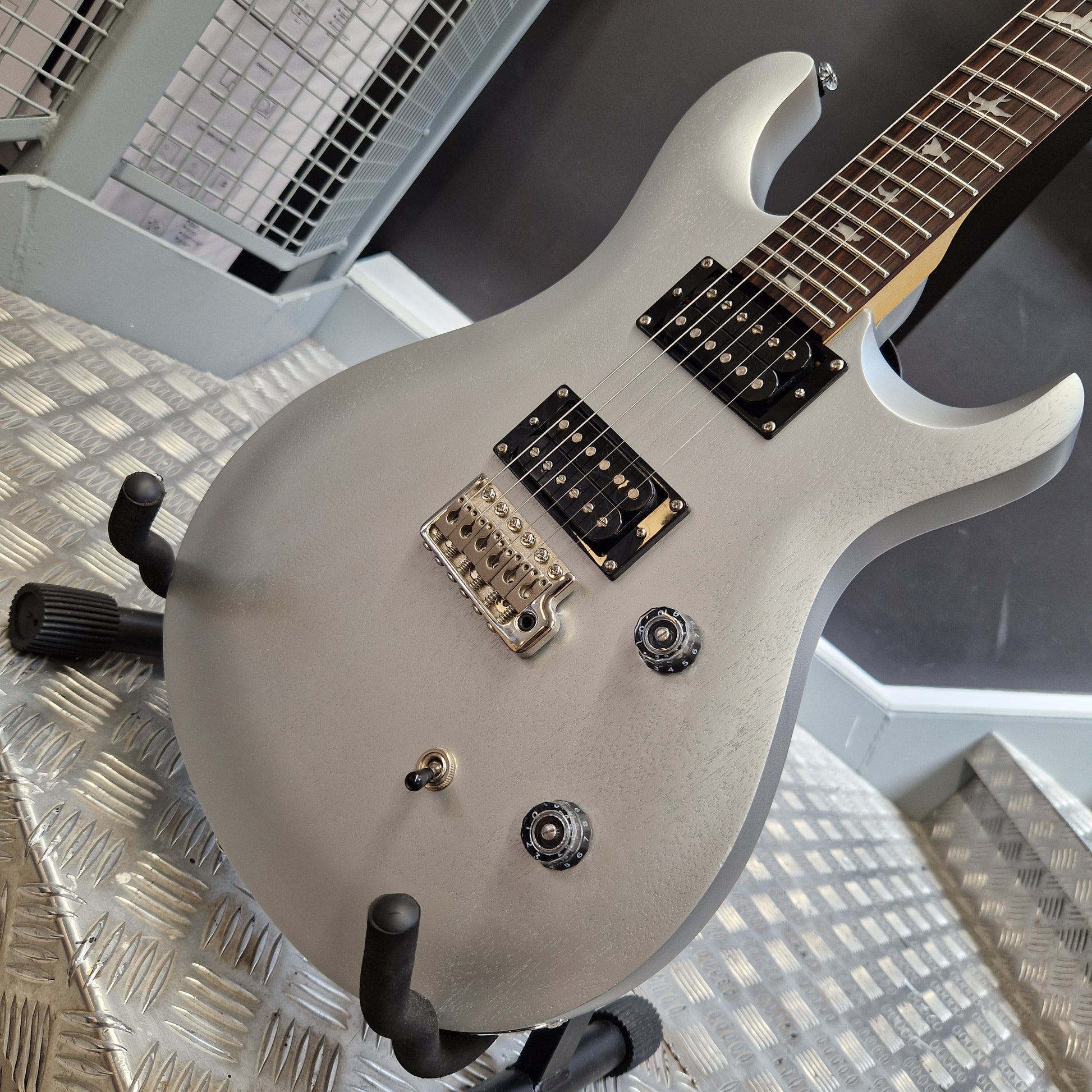 Pre-Owned PRS SE CE24 A close-up of an electric guitar body in a matte silver finish, mounted on a stand. The body features black hardware, a gold-toned tremolo bridge, and a light wood neck visible at the top left, set against a metallic diamond plate floor.