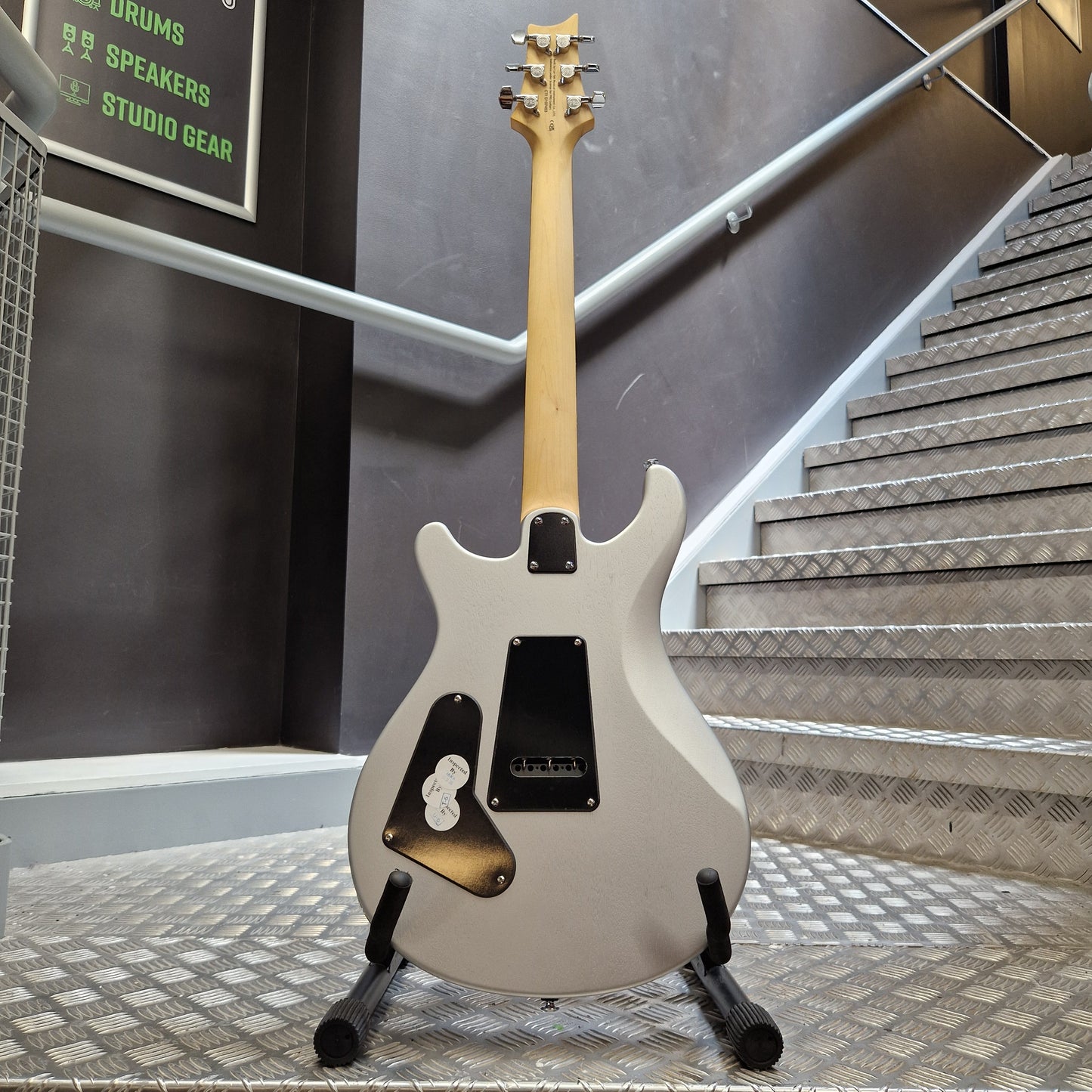 Pre-Owned PRS SE CE24 A white electric guitar with a natural wood neck and black pickups, resting on a stand in a metal workshop environment.