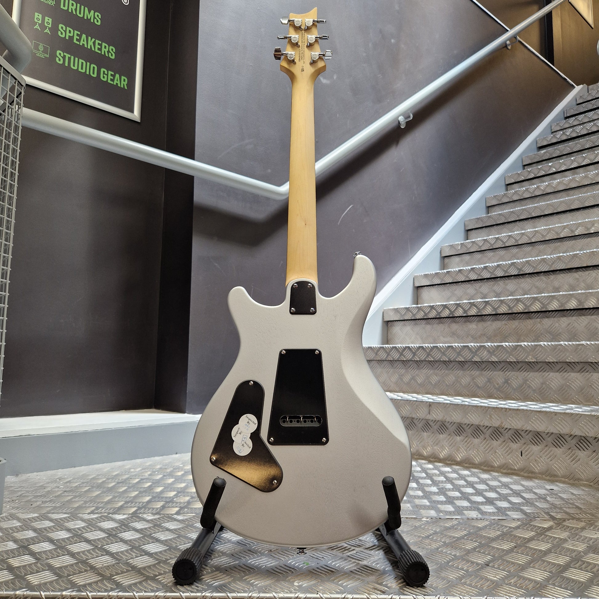 Pre-Owned PRS SE CE24 A white electric guitar with a natural wood neck and black pickups, resting on a stand in a metal workshop environment.