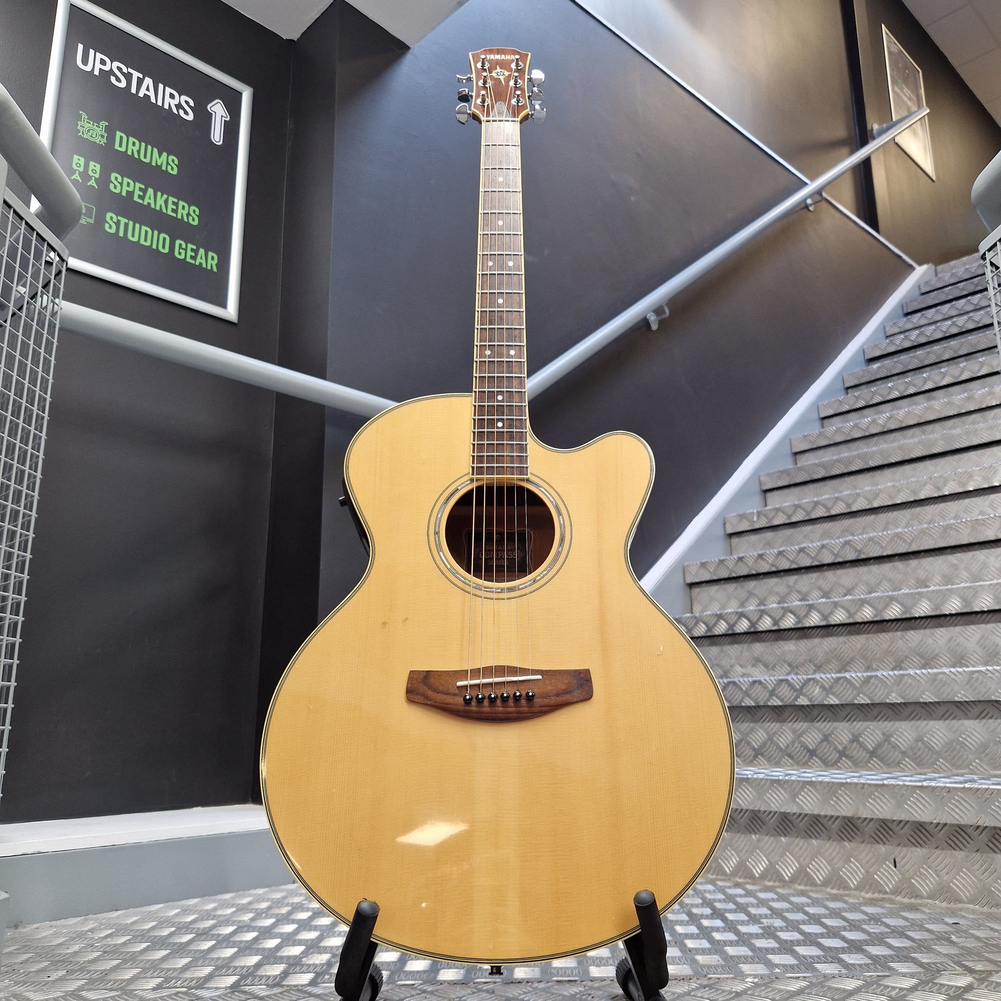 Pre-Owned Yamaha CPX700 A natural wood cutaway acoustic-electric guitar hanging on a display stand in a studio or showroom with dark walls and metal paneling.