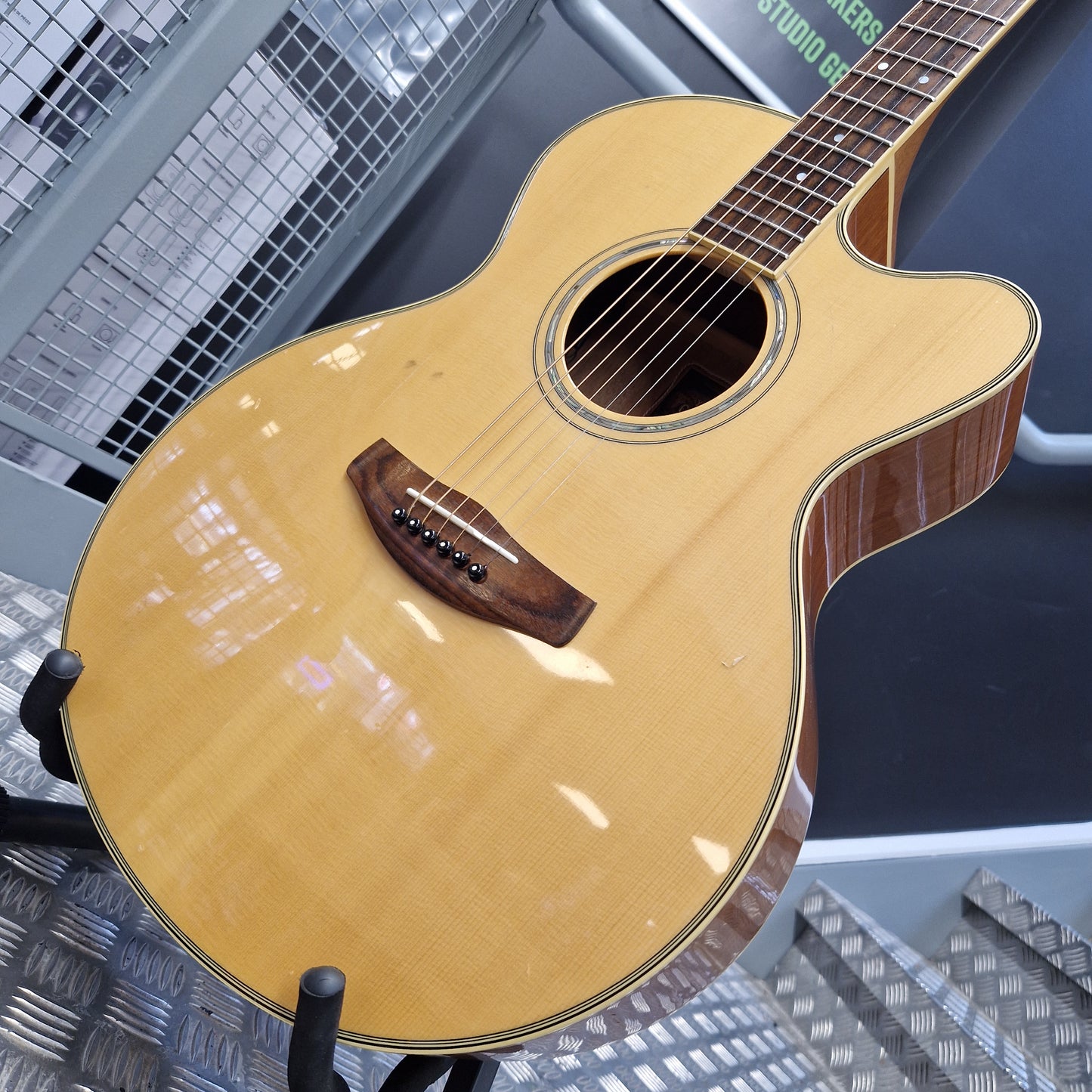 Pre-Owned Yamaha CPX700 A close-up of a Taylor acoustic guitar with a natural wood finish and dark wood details, resting on a stand in what appears to be a showroom or studio.