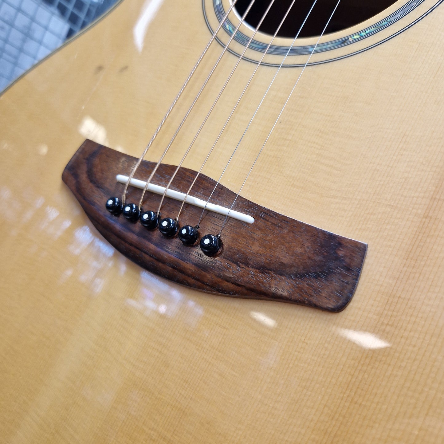 Pre-Owned Yamaha CPX700 A close-up of an acoustic guitar body showing a dark brown wooden bridge with six black saddle-style tuners and silver strings over a natural wood grain face.