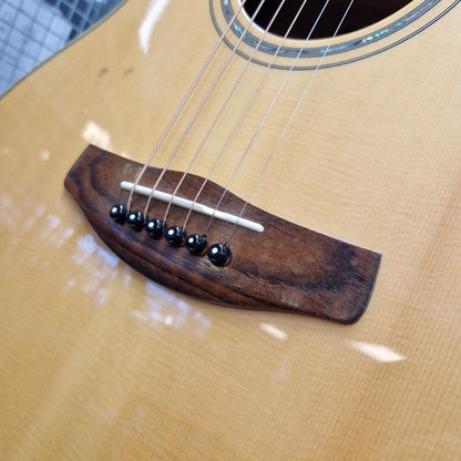 Pre-Owned Yamaha CPX700 A close-up of an acoustic guitar body showing a dark brown wooden bridge with six black saddle-style tuners and silver strings over a natural wood grain face.