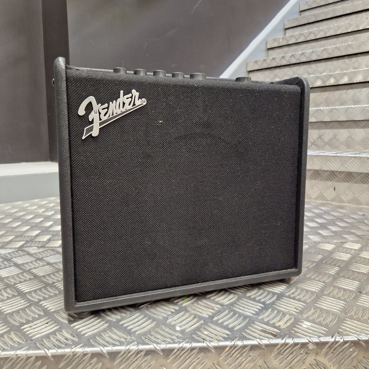 Pre-Owned Fender A Fender guitar amplifier speaker cabinet with a black textured fabric grille and the silver Fender logo, photographed on a diamond-plate metal surface.