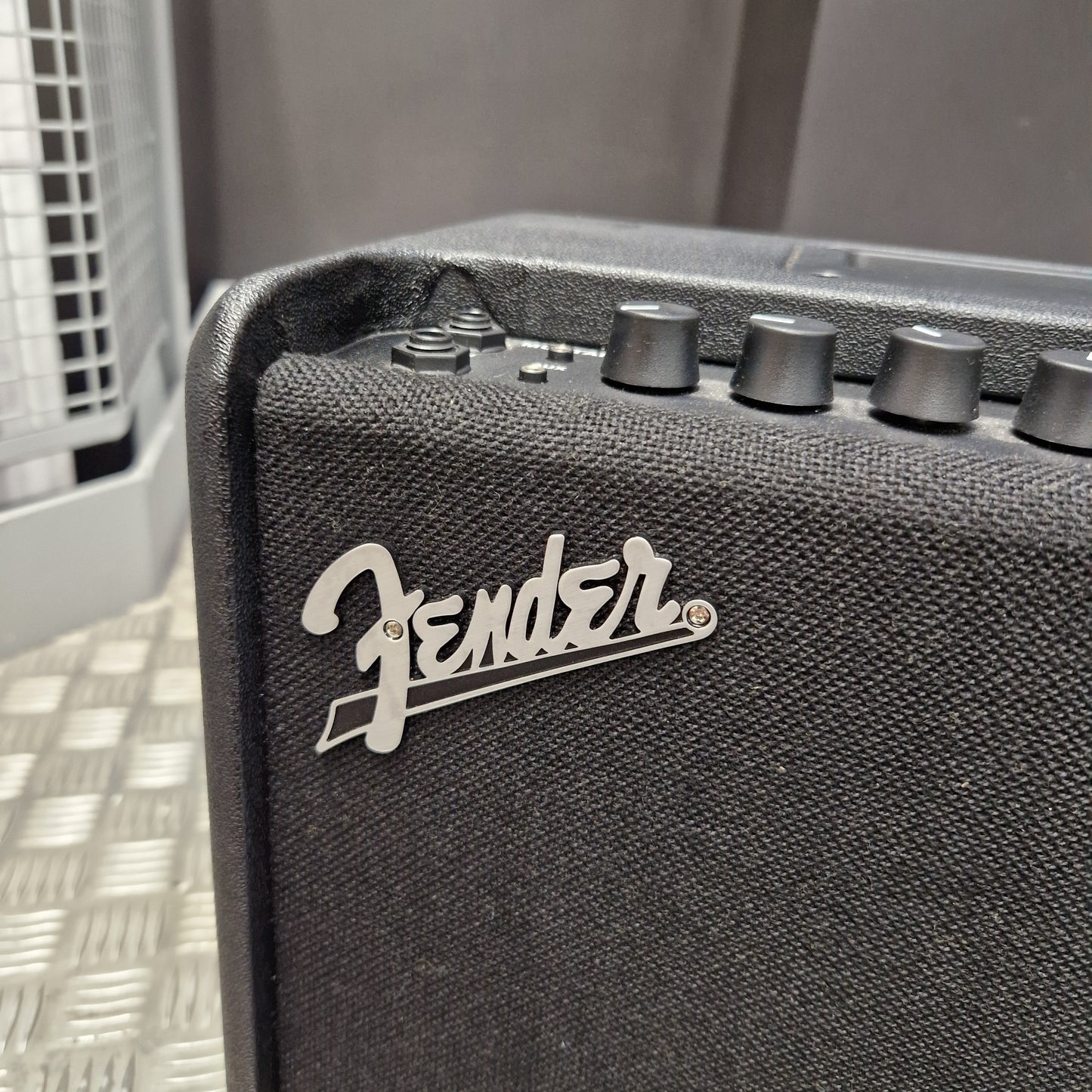Pre-Owned Fender Close-up of a black Fender guitar amplifier corner showing the silver Fender logo and control knobs against a dark textured fabric grille.