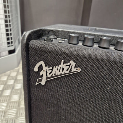 Pre-Owned Fender Close-up of a black Fender guitar amplifier corner showing the silver Fender logo and control knobs against a dark textured fabric grille.