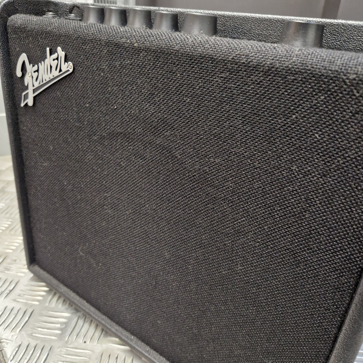 Pre-Owned Fender Close-up of a black Fender speaker grille with the silver Fender logo in the lower-left corner, set against a silver diamond plate background.