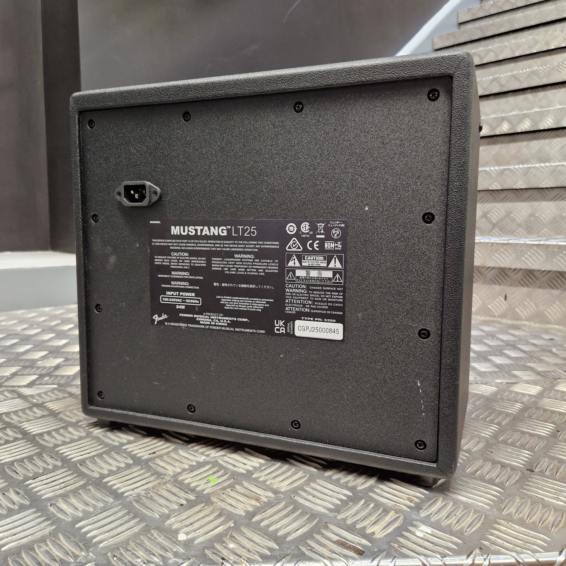 Pre-Owned Fender The underside of a black Mustang LT25 speaker cabinet, showing the mounting holes, power connector, and product specifications label, resting on a diamond plate metal surface.