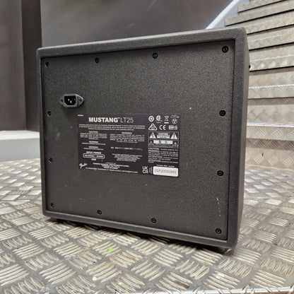 Pre-Owned Fender The underside of a black Mustang LT25 speaker cabinet, showing the mounting holes, power connector, and product specifications label, resting on a diamond plate metal surface.