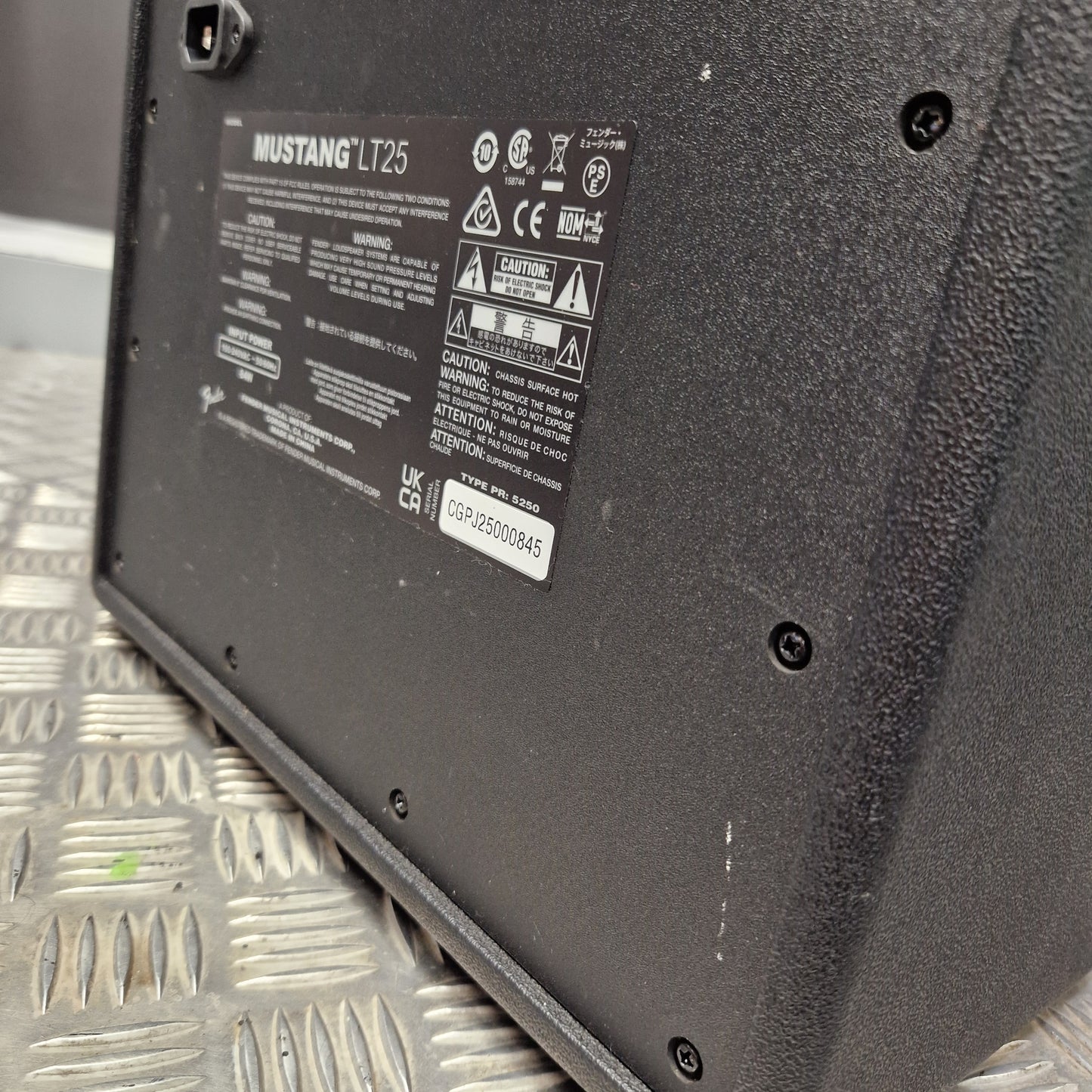 Pre-Owned Fender Close-up of the underside of a black metal equipment housing or panel, showing screws, a product label, and a power cord connector, resting on a silver diamond plate.