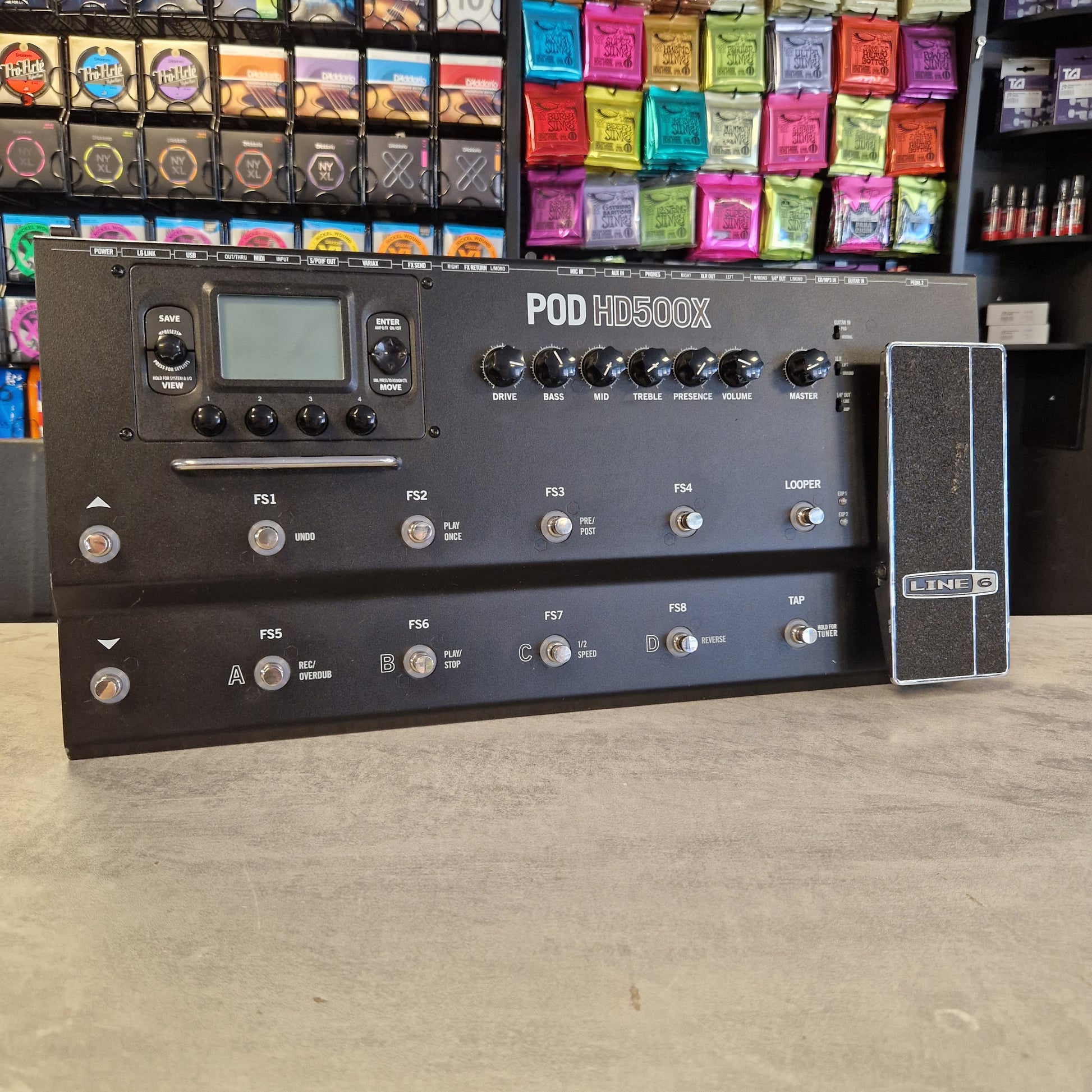 Pre-Owned Line 6 A black Fuzz Face 100 FX guitar effects pedal with multiple knobs, a small display screen, and footswitches, photographed in a store setting with shelves of colorful guitar pedals visible on the left.