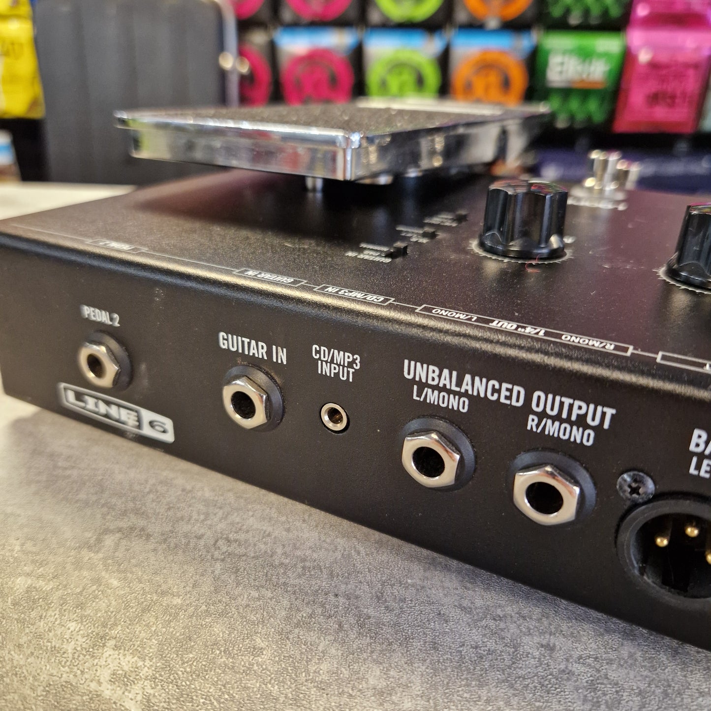 Pre-Owned Line 6 Close-up of the rear panel of a black audio device, showing jacks labeled unbalanced output, guitar in, and other audio inputs, with knobs and a pedal visible.