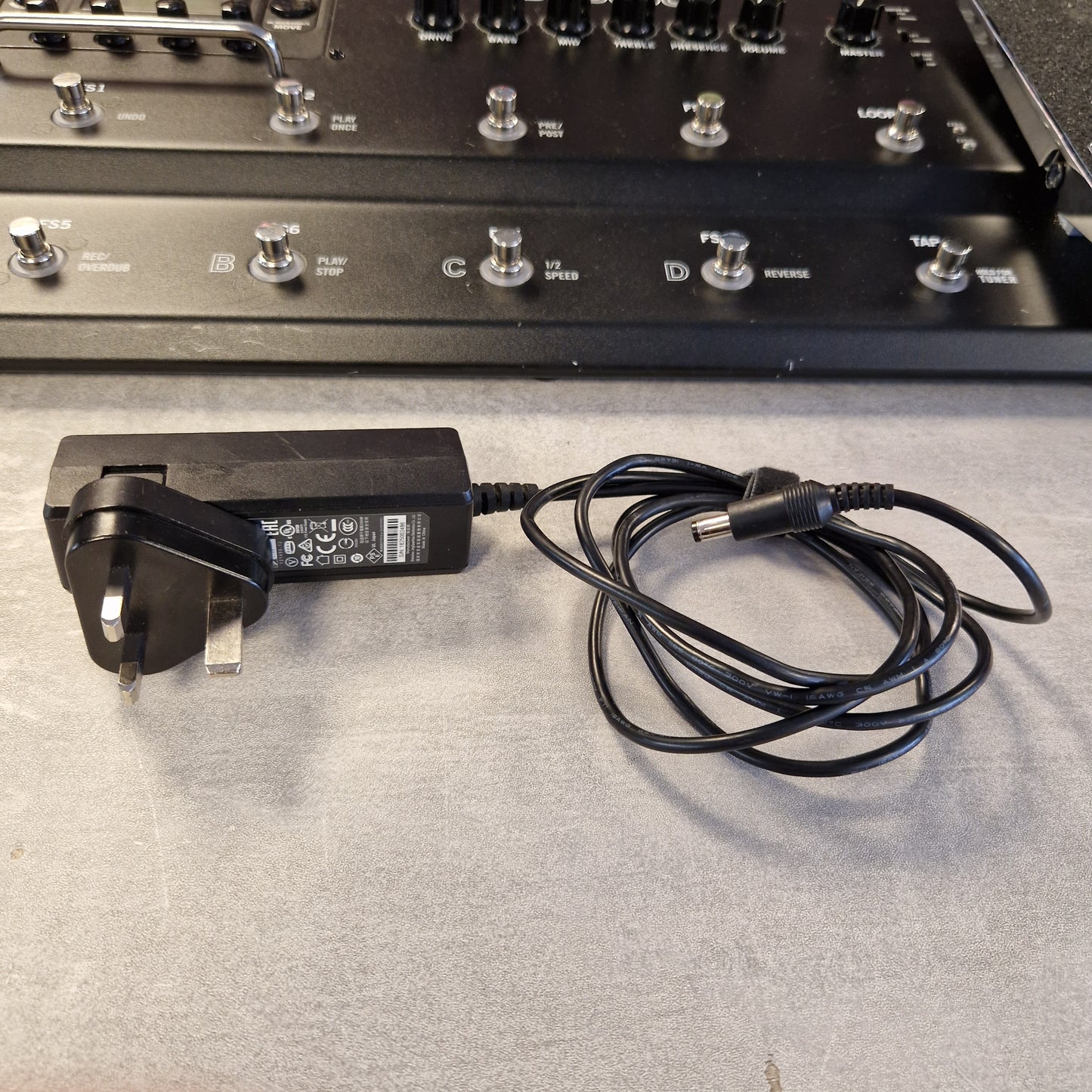 Pre-Owned Line 6 A black AC adapter power supply with a coiled black cable and UK two-pin plug, photographed next to a dark electronic control panel on a gray surface.