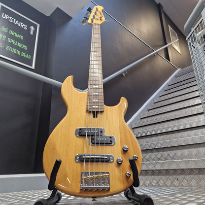 Pre-Owned Yamaha BB615 A natural wood electric bass guitar on a stand in an indoor stairwell area, with a dark wall, metal handrails, and a sign reading 'UPSTAIRS' in the background.