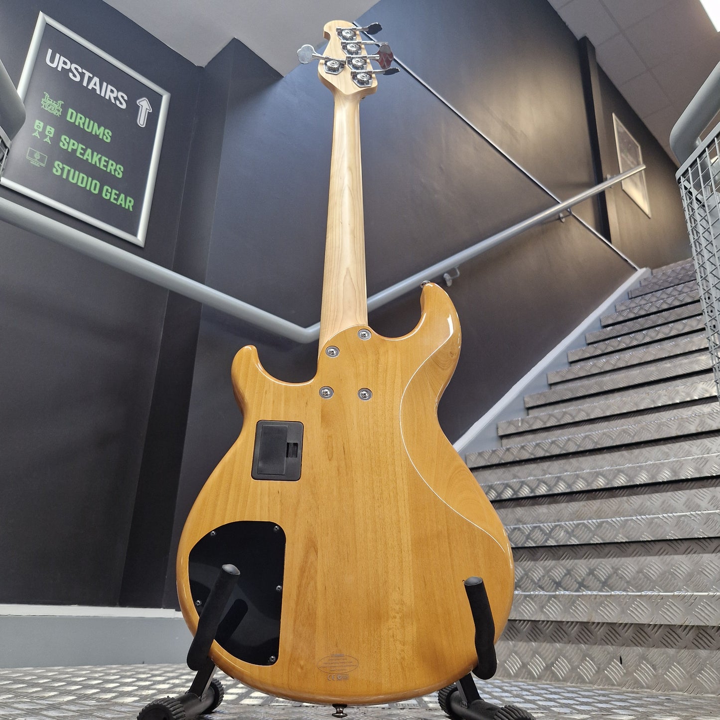 Pre-Owned Yamaha BB615 A natural wood electric bass guitar shown from the back, resting on a stand in an indoor stairwell area.