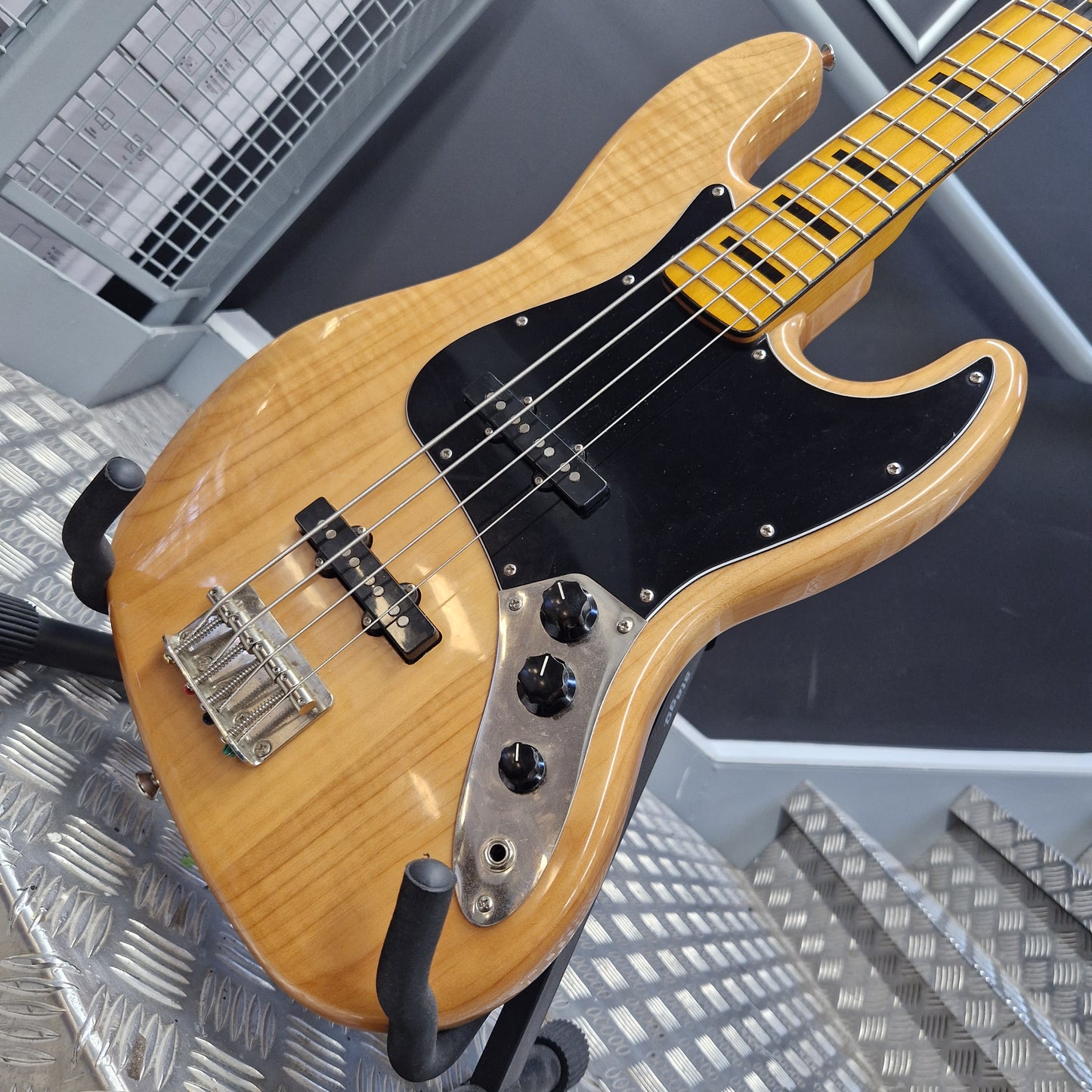 Pre-Owned Squier  A four-string bass guitar with a natural maple wood finish, black pickguard, and chrome hardware, resting on a stand in a workshop setting.