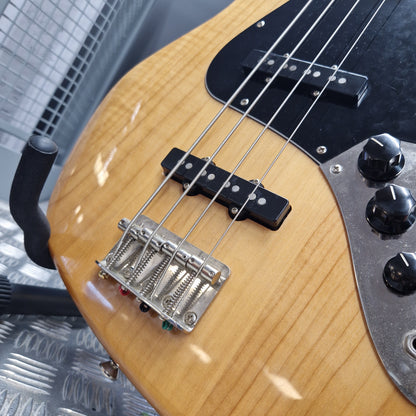 Pre-Owned Squier  Close-up of a natural wood four-string bass guitar showing the body, black pickguard, pickups, bridge, and part of the control knobs.