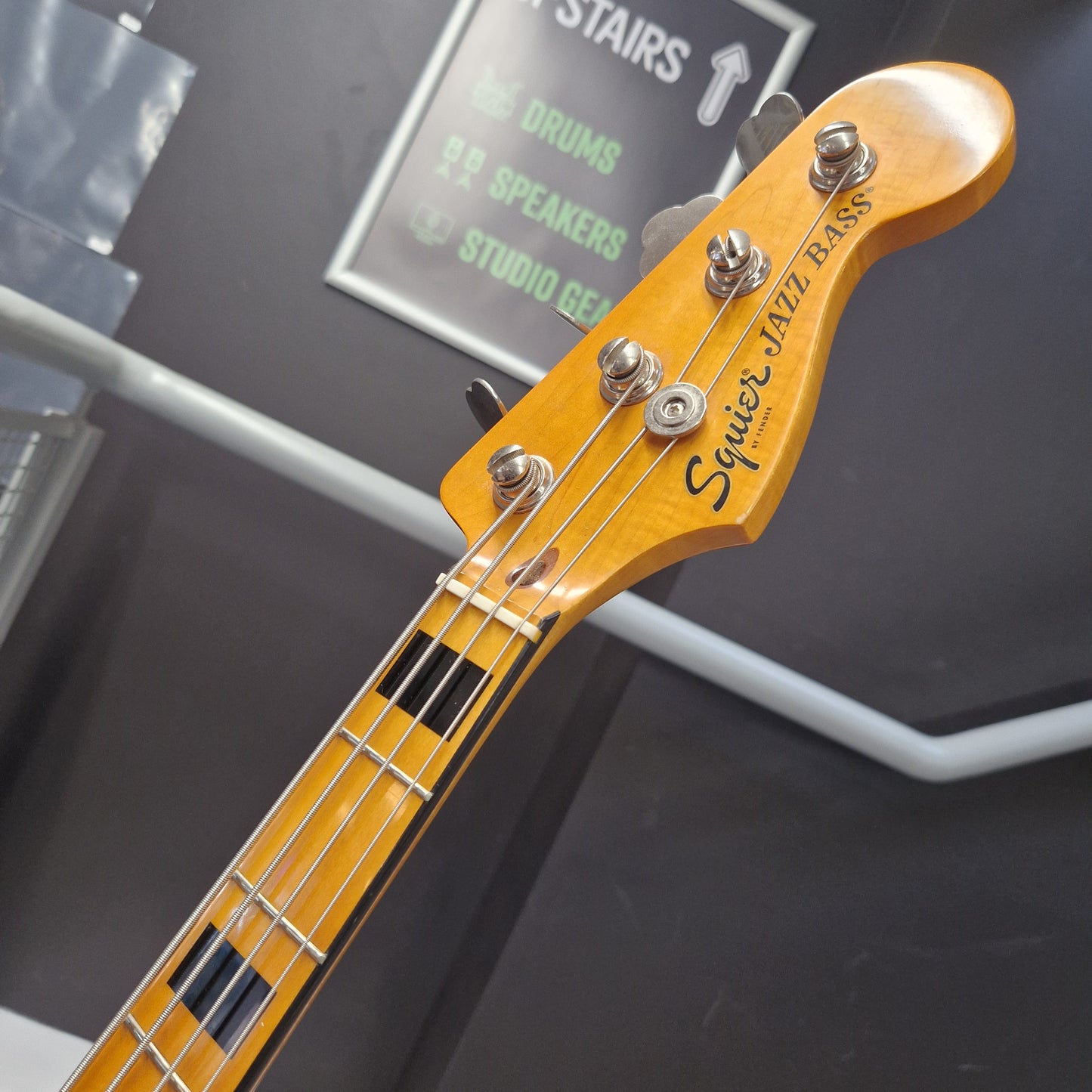 Pre-Owned Squier  Close-up of a Squier Jazz Bass headstock and upper neck, showing the yellow-orange wood finish, four silver tuning pegs, and black fretboard block inlays against a dark gray background.
