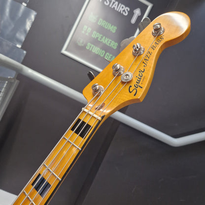 Pre-Owned Squier  Close-up of a Squier Jazz Bass headstock and upper neck, showing the yellow-orange wood finish, four silver tuning pegs, and black fretboard block inlays against a dark gray background.