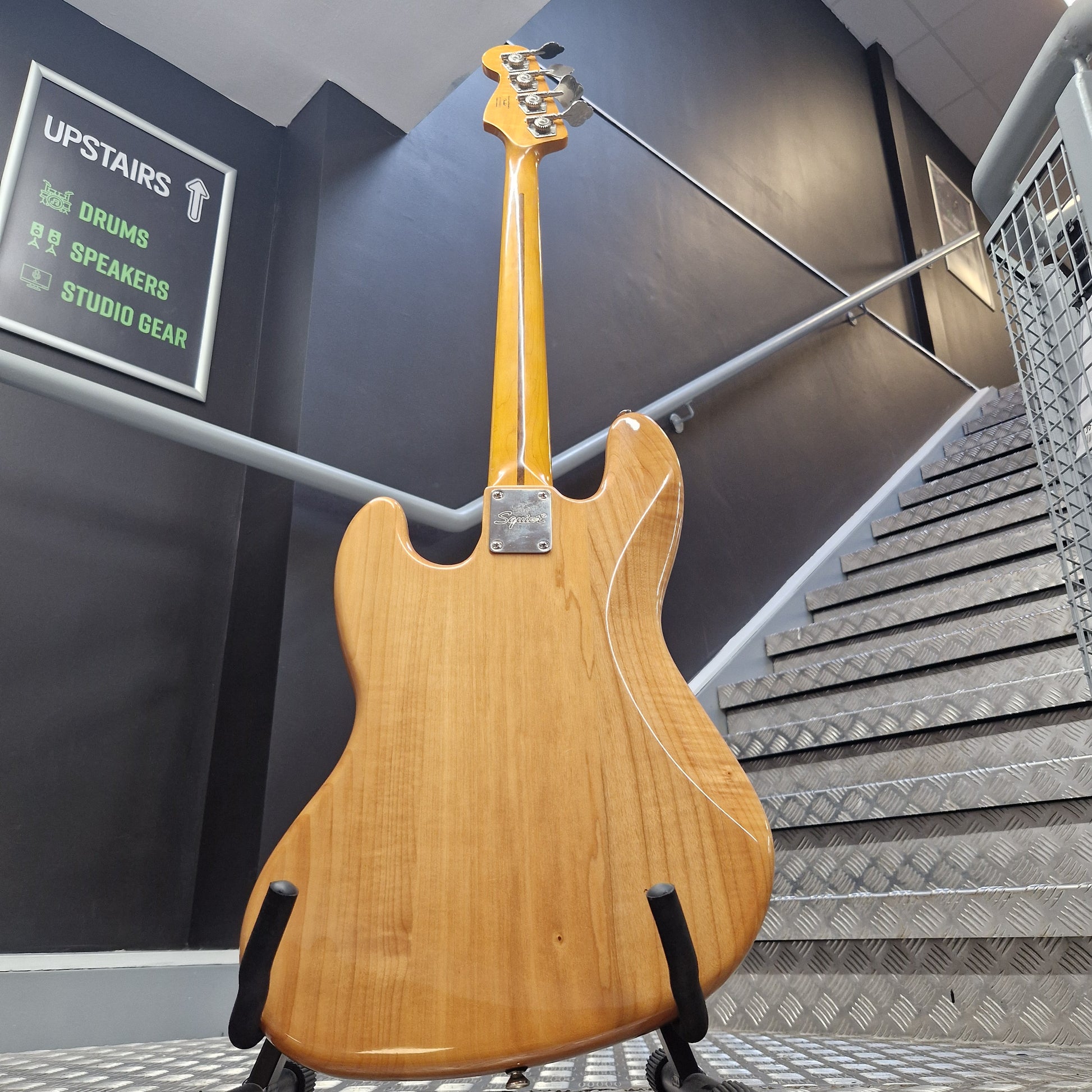 Pre-Owned Squier  A natural wood electric guitar body and neck are shown hanging from black support straps in an indoor space with a metal grate wall and dark floor.