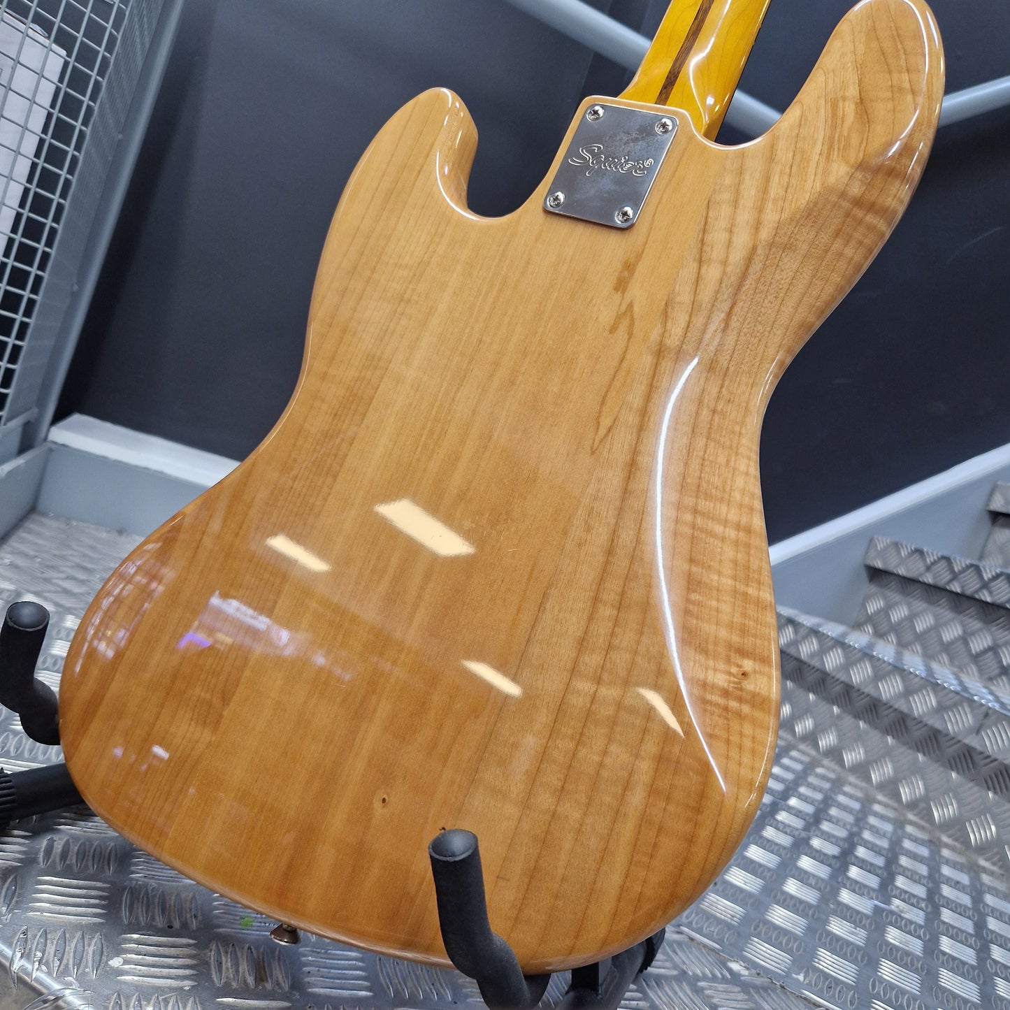 Pre-Owned Squier  Close-up of the back of an electric guitar body with a glossy natural wood finish, showing the grain pattern and a black pickguard on a metal grid surface.