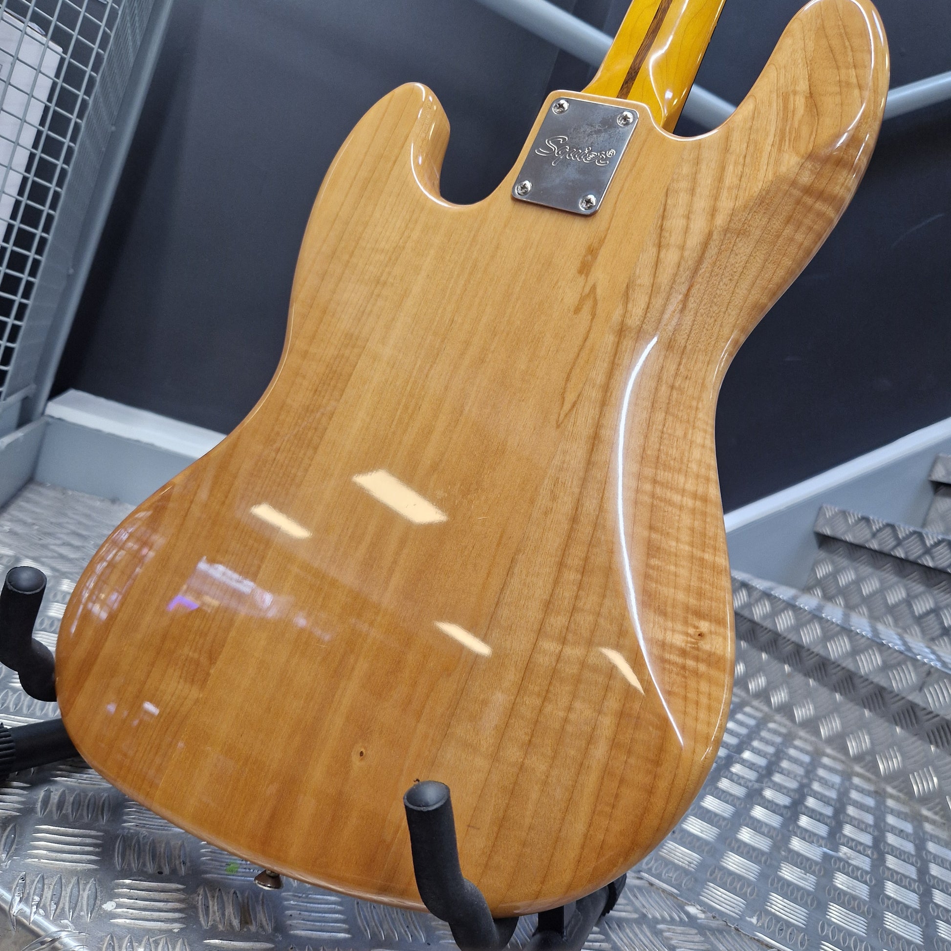 Pre-Owned Squier  Close-up of the back of an electric guitar body with a glossy natural wood finish, showing the grain pattern and a black pickguard on a metal grid surface.