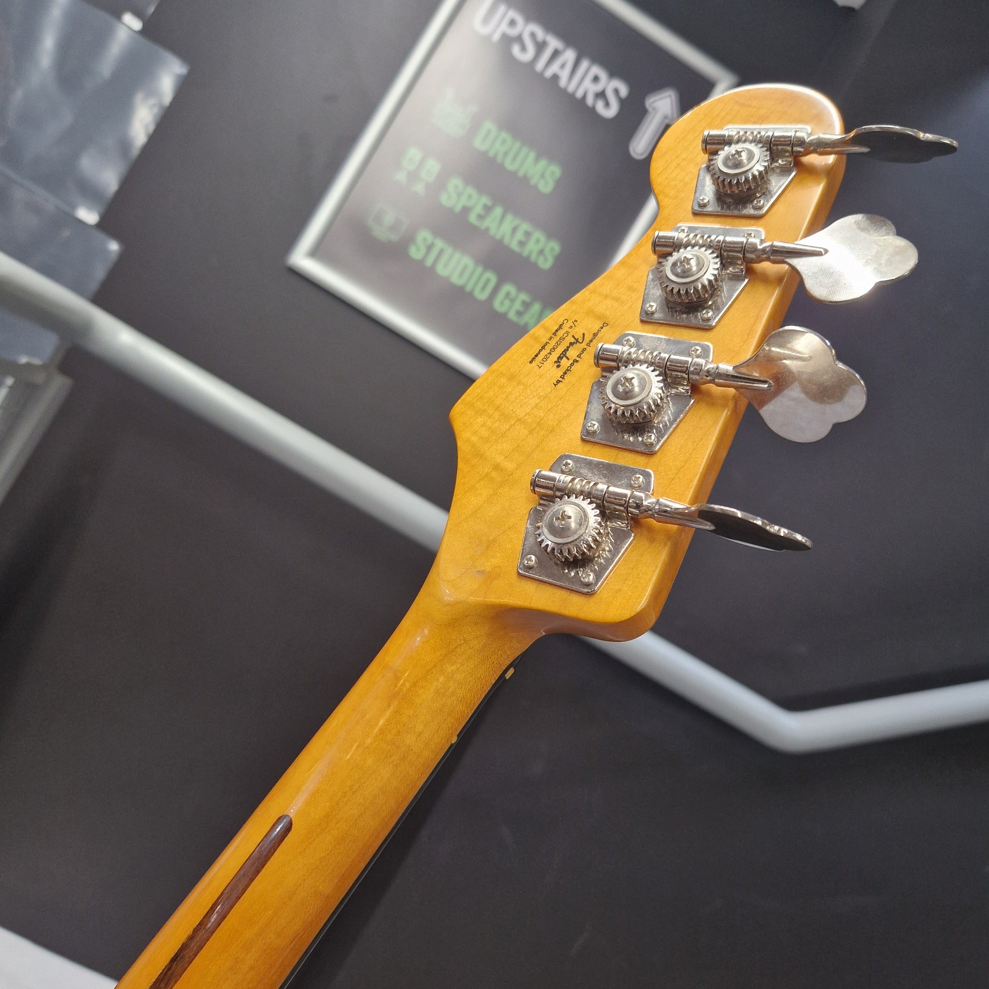 Pre-Owned Squier  Close-up of a four-string bass guitar headstock with a natural maple wood finish and silver tuning pegs, photographed indoors against a dark gray background.
