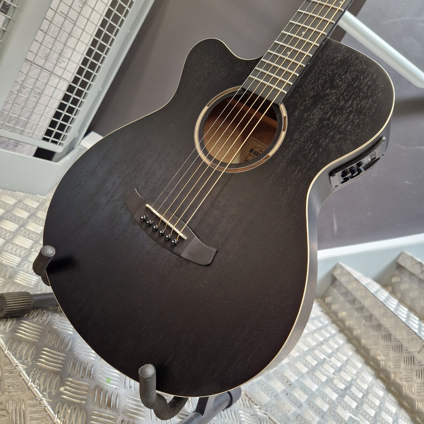 Pre-Owned Tanglewood TWBBSFCELH A black acoustic-electric guitar with a glossy finish and cutaway body, resting on a stand in an indoor setting.