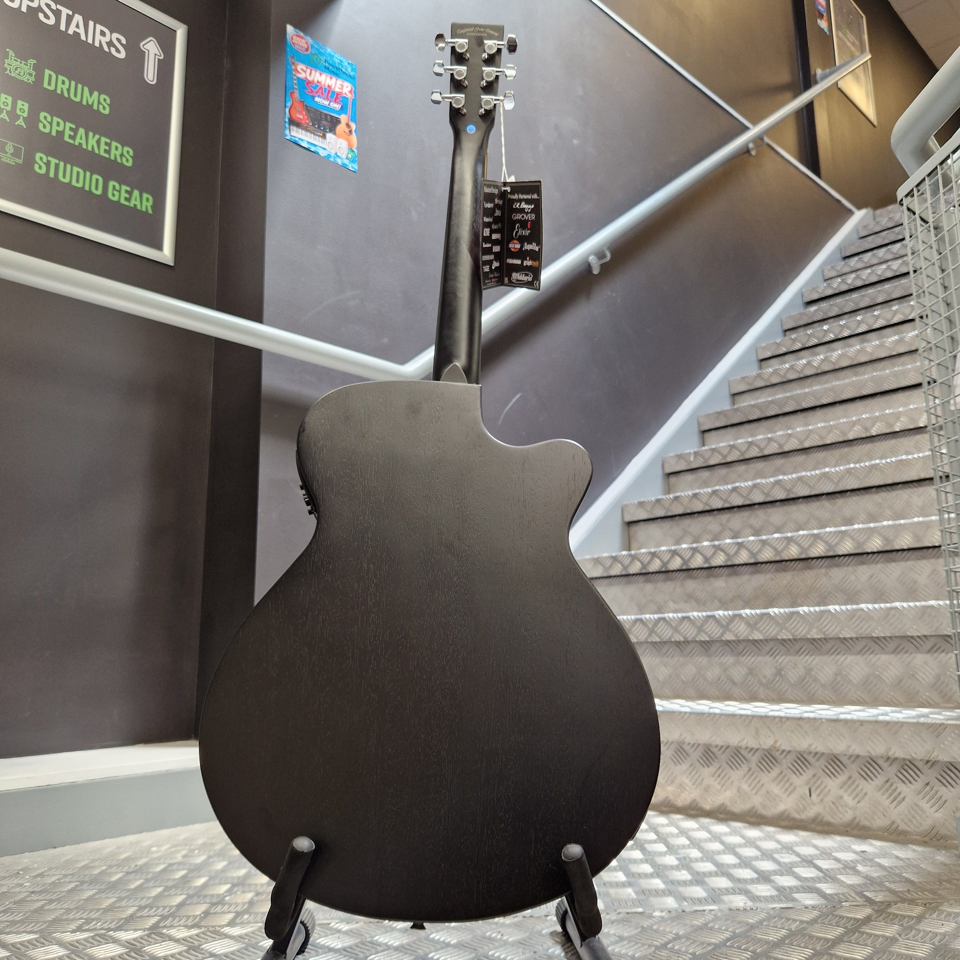 Pre-Owned Tanglewood TWBBSFCELH A black electric guitar body and neck are shown hanging on a display stand inside a store or showroom, with metal shelving and a sign visible in the background.