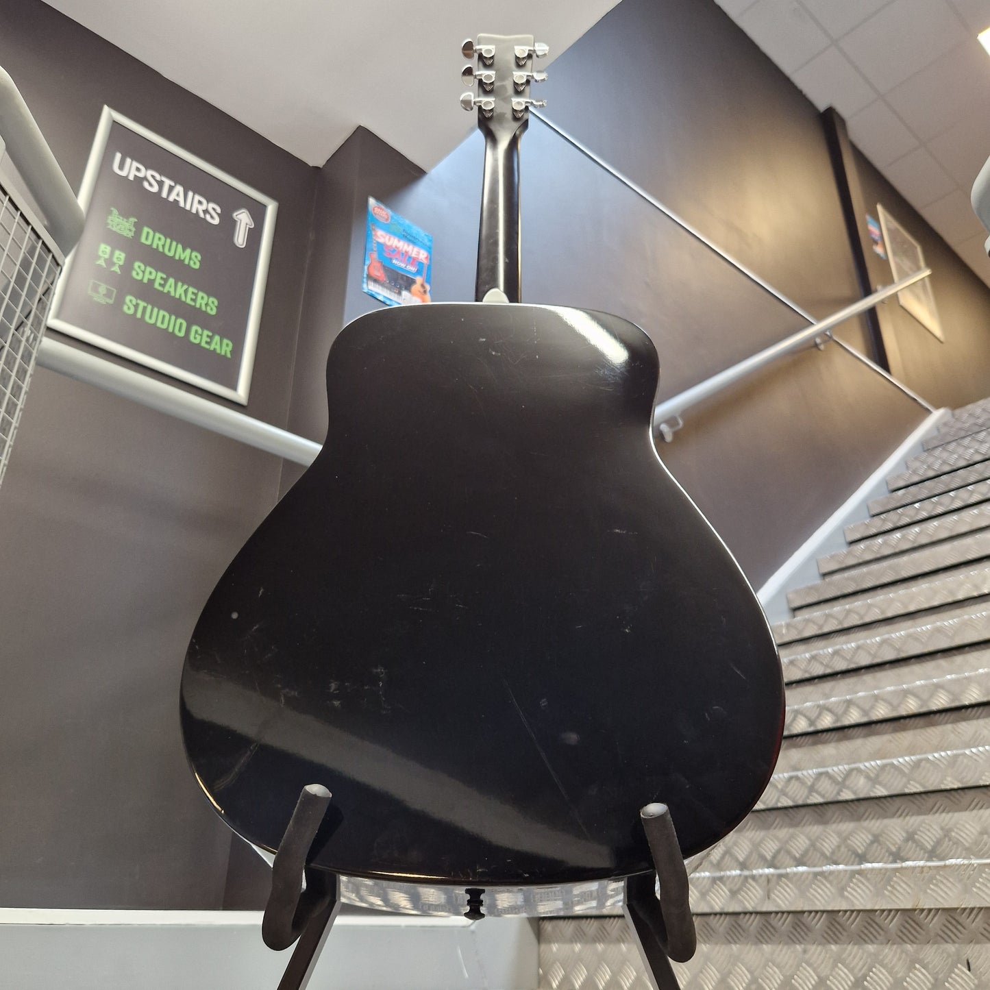 Pre-Owned Yamaha FG800 A black electric guitar body and neck are shown hanging on a display stand inside a store or showroom, with store fixtures, signage, and metal paneling visible in the background.