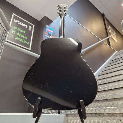 Pre-Owned Yamaha FG800 A black electric guitar body and neck are shown hanging on a display stand inside a store or showroom, with store fixtures, signage, and metal paneling visible in the background.