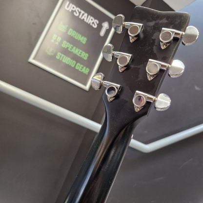 Pre-Owned Yamaha FG800 Close-up of the headstock of a black electric guitar, showing six silver tuning pegs, with a blurred background containing a sign that reads 'UPSTAIRS' and 'STUDIO GEAR'.