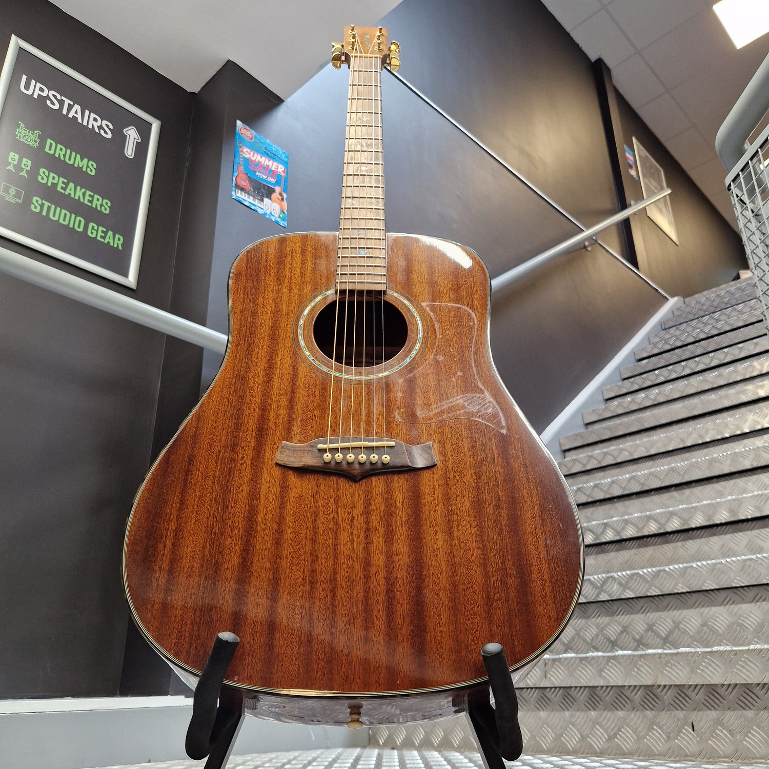 Pre-Owned Tanglewood TW15ASMNAT A wooden acoustic guitar with a glossy brown finish stands on a black display stand inside a store or studio setting, with stairs and signage visible in the background.