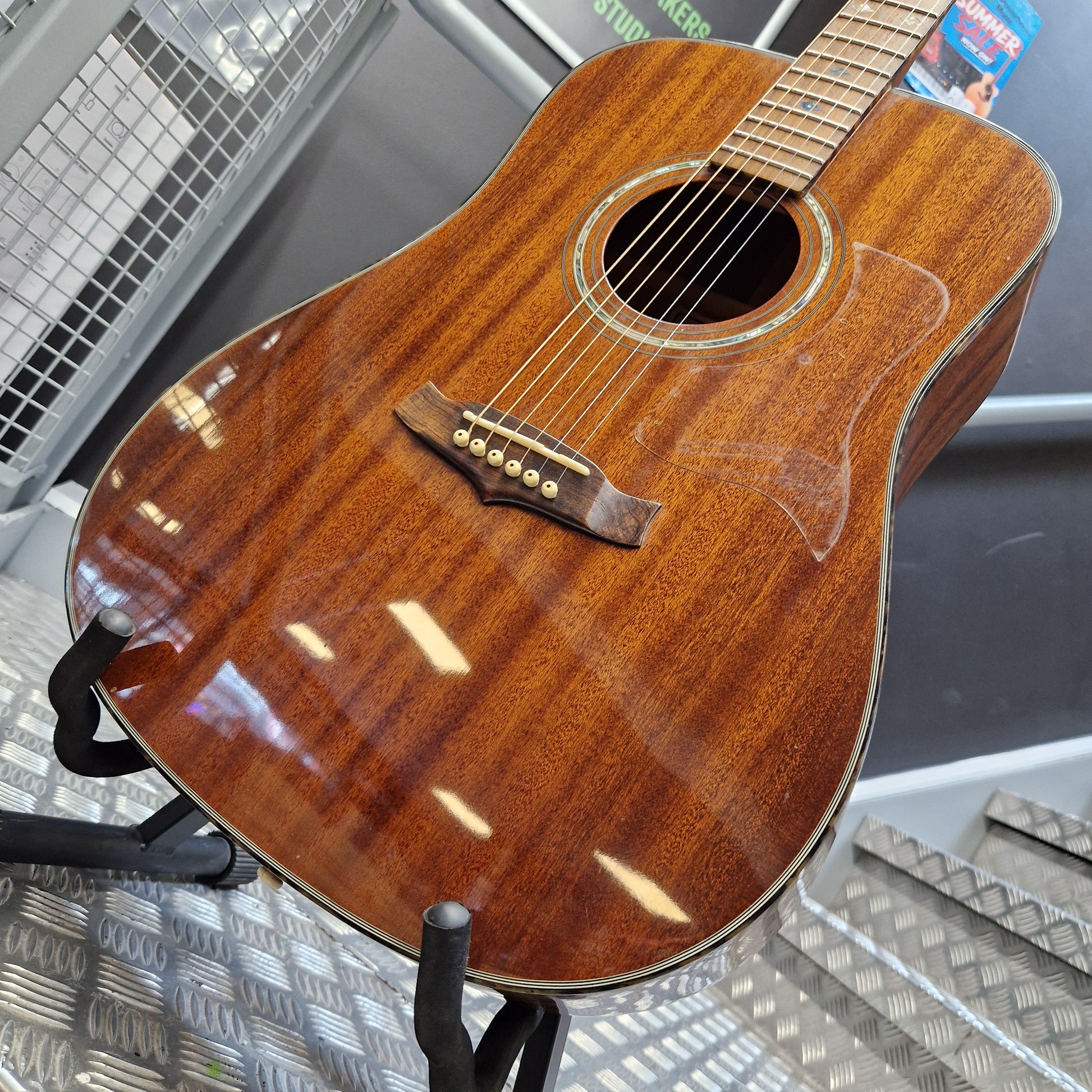 Pre-Owned Tanglewood TW15ASMNAT A close-up of a wooden acoustic guitar body with a glossy finish, showing the sound hole, pickguard, and bridge, resting on a stand in what appears to be a store or workshop.