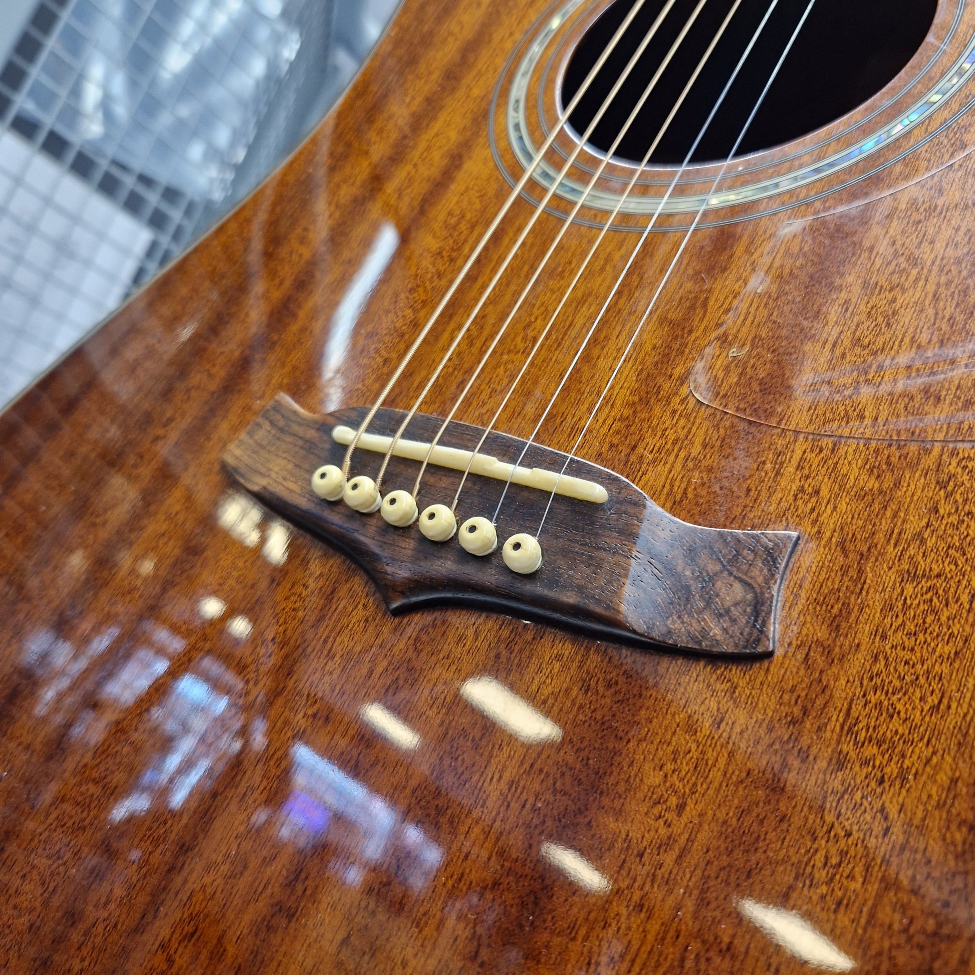 Pre-Owned Tanglewood TW15ASMNAT Close-up of an acoustic guitar body showing the brown wooden bridge with cream-colored saddle and tuning peg bars, set against a glossy wood finish with reflections.
