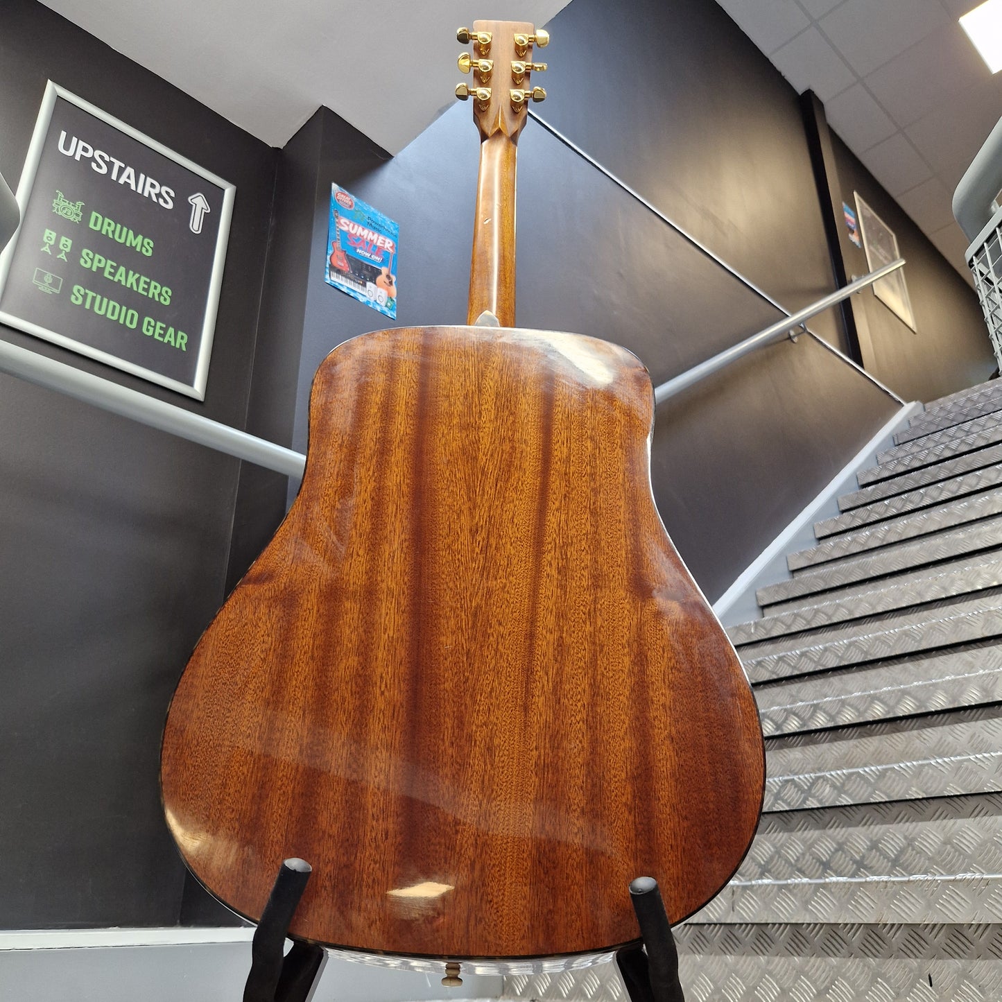 Pre-Owned Tanglewood TW15ASMNAT A wooden acoustic guitar shown from the back, resting on a black stand in a real indoor environment with dark walls, a staircase, and signage in the background.
