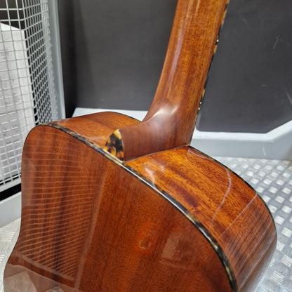 Pre-Owned Tanglewood TW15ASMNAT Close-up of the back of a glossy brown acoustic guitar, showing the wood grain, sound hole, and tortoiseshell-style binding against a gray and white industrial background.