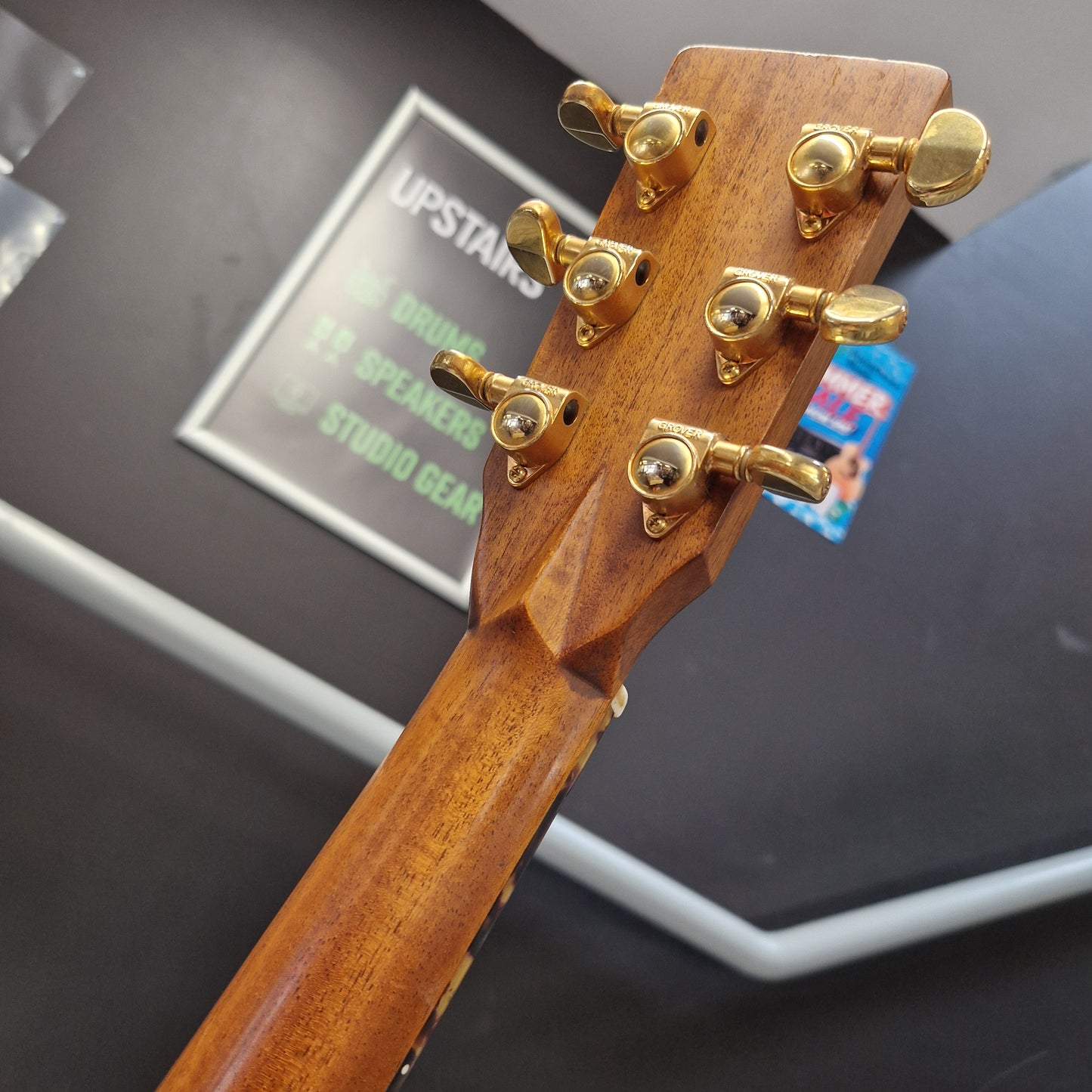 Pre-Owned Tanglewood TW15ASMNAT Close-up of the headstock of a wooden drum kit, showing six gold metal tuning pegs on a dark wood neck, with a blurred background containing a sign that reads 'UPSTARS'.