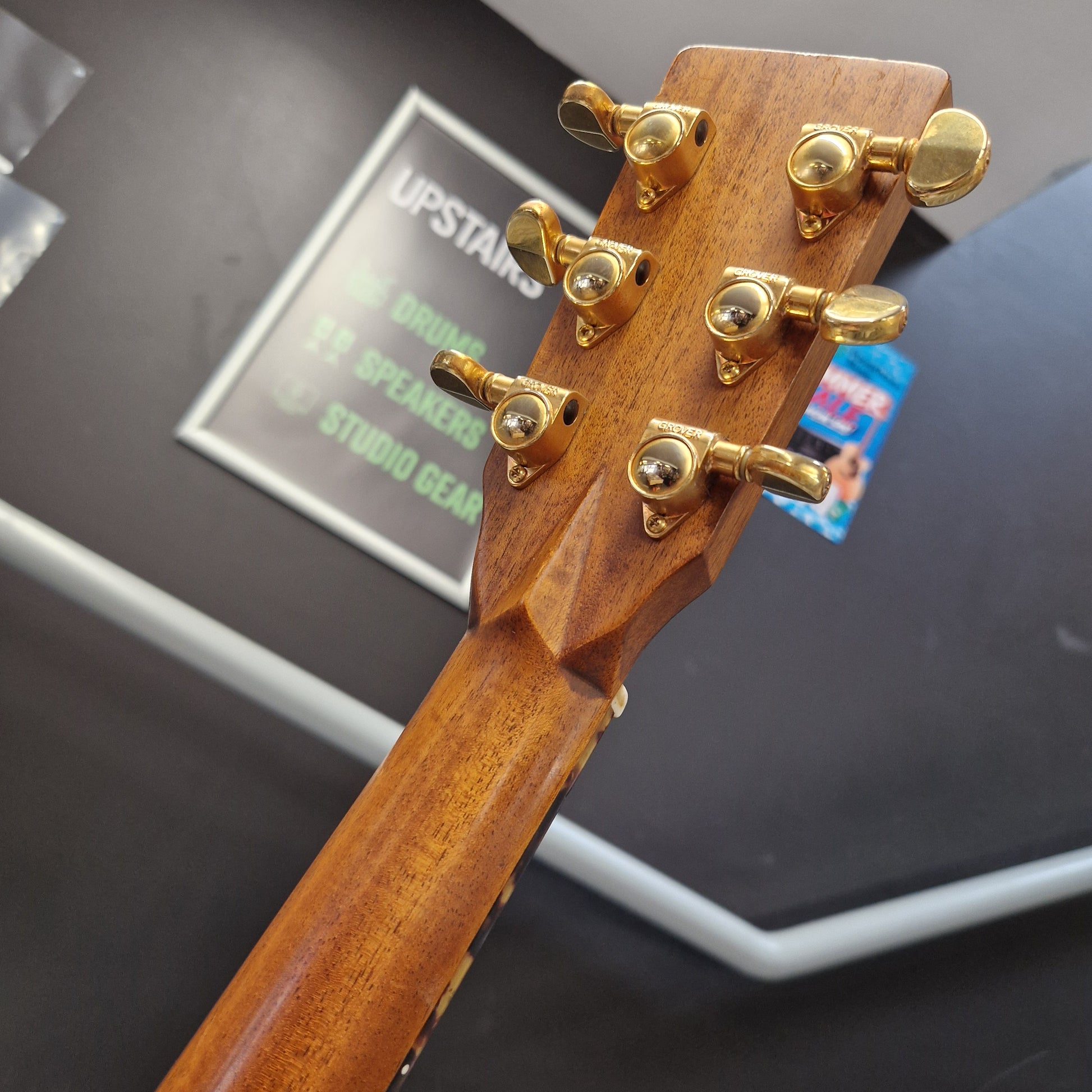 Pre-Owned Tanglewood TW15ASMNAT Close-up of the headstock of a wooden drum kit, showing six gold metal tuning pegs on a dark wood neck, with a blurred background containing a sign that reads 'UPSTARS'.