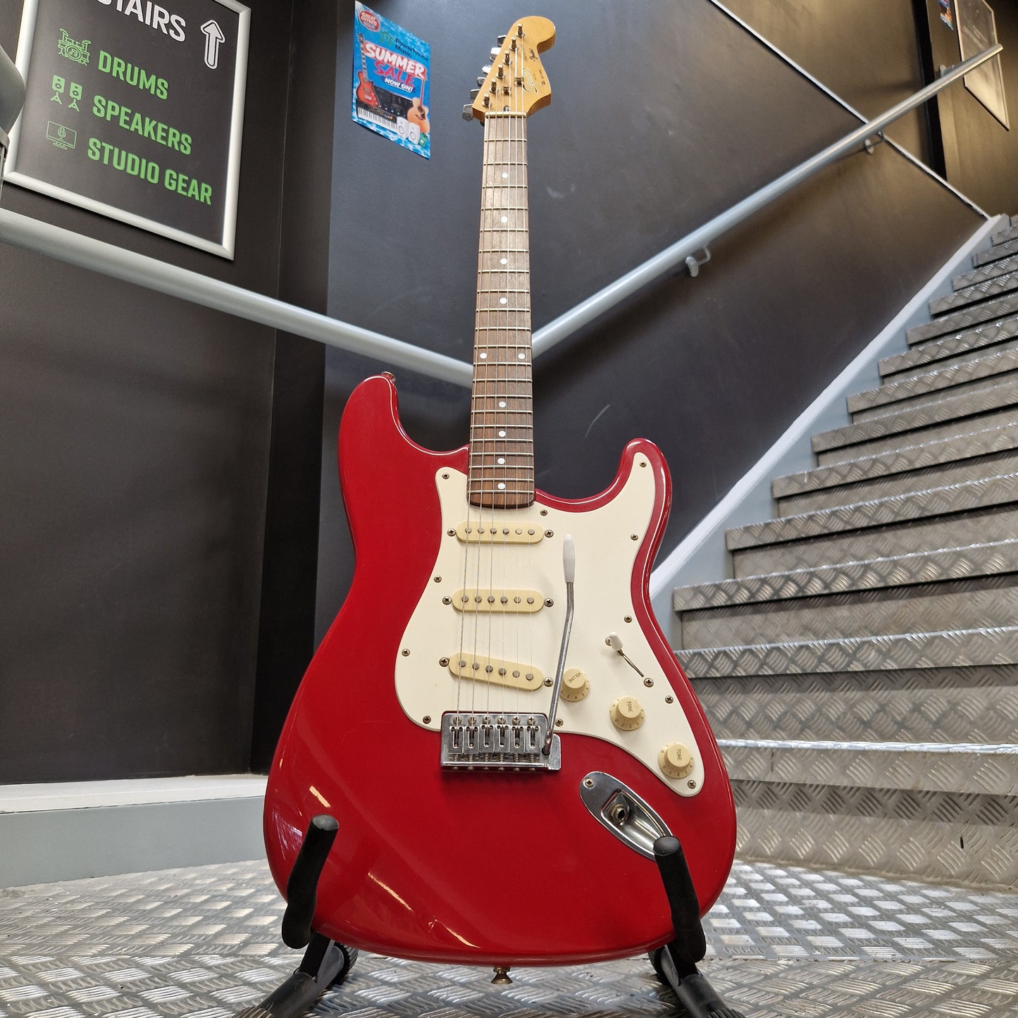 Pre-Owned Squier MIK 1992 A red electric guitar with a cream pickguard and gold-toned controls, displayed on a stand in an indoor showroom with metal wall panels and a dark floor.