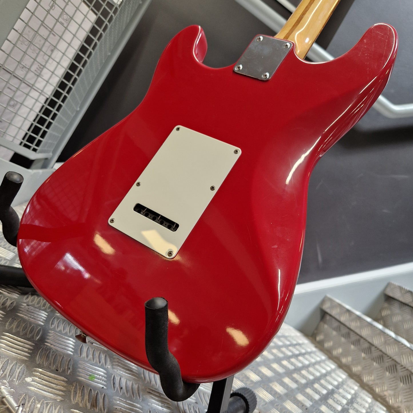 Pre-Owned Squier MIK 1992 A red electric guitar body shown from the back, mounted on a black stand in an industrial setting with metal grates visible in the background.