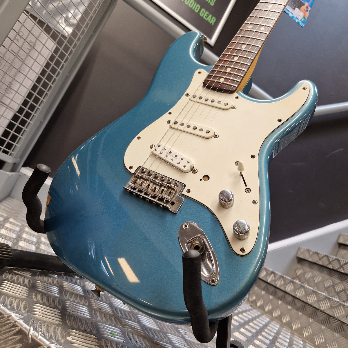 Pre-Owned Fender Mexican Stratocaster 1993 Electric - Lake Placid Blue A blue electric guitar with a cream pickguard and chrome hardware, photographed indoors on a stand with metal wall panels in the background.