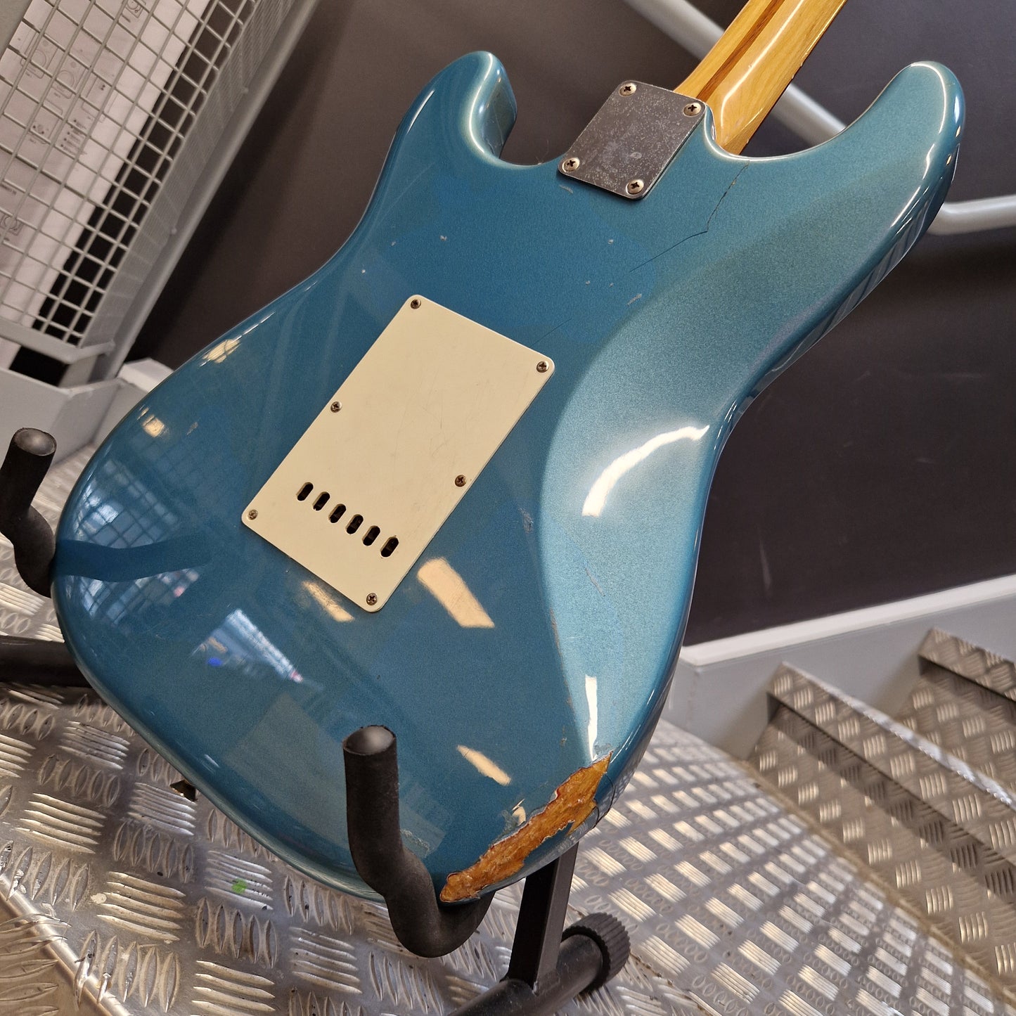 Pre-Owned Fender Mexican Stratocaster 1993 Electric - Lake Placid Blue Close-up of the back of a blue electric guitar resting on a stand, showing a cream pickguard, a worn orange patch on the body, and part of a light wood neck.