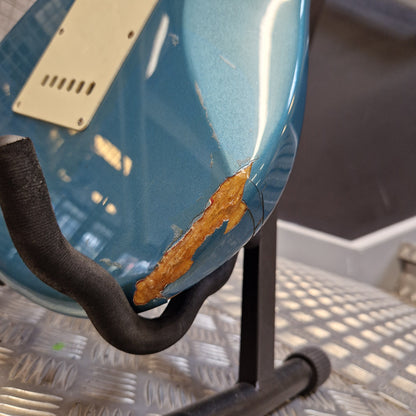 Pre-Owned Fender Mexican Stratocaster 1993 Electric - Lake Placid Blue Close-up of a blue electric guitar body with a prominent scratch and rusted paint damage on the side, resting on a metal grate surface.
