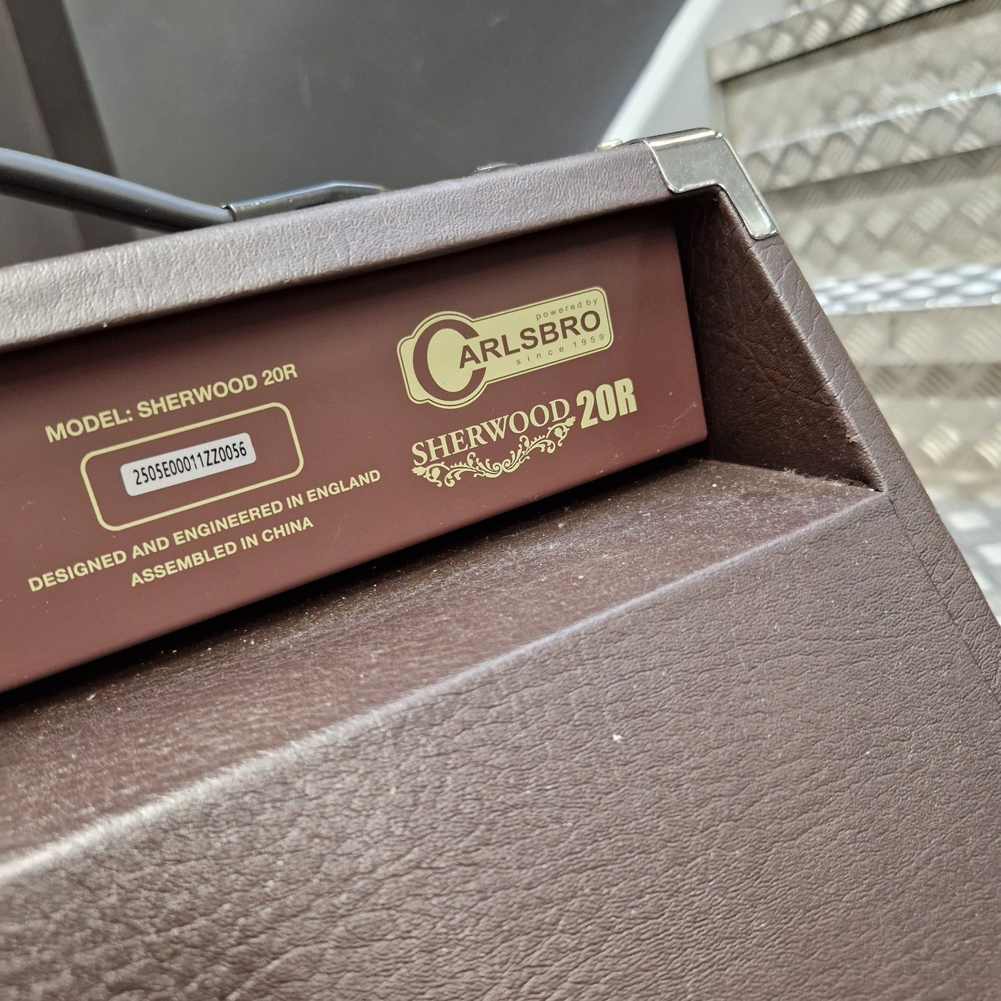Pre-Owned Carlsbro Sherwood 20R A close-up of a brown textured box containing a dark brown cardboard insert labeled 'CARLSBRO SHENWOOD 20R' with gold text and a barcode, placed on a dark surface with a patterned curtain in the background.