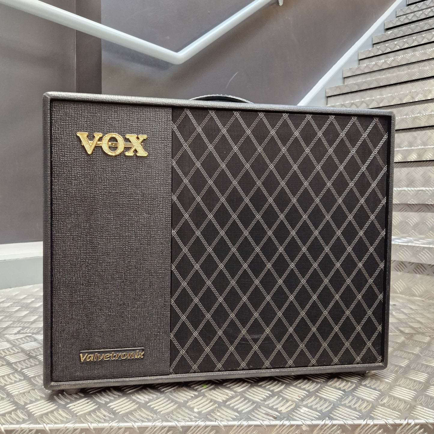 Pre-Owned Vox VT100X A Vox Valetcronix guitar amplifier speaker cabinet with a dark gray textured grille and a black diamond-patterned front, featuring gold 'VOX' lettering, placed on a metal diamond-plate surface with stairs in the background.
